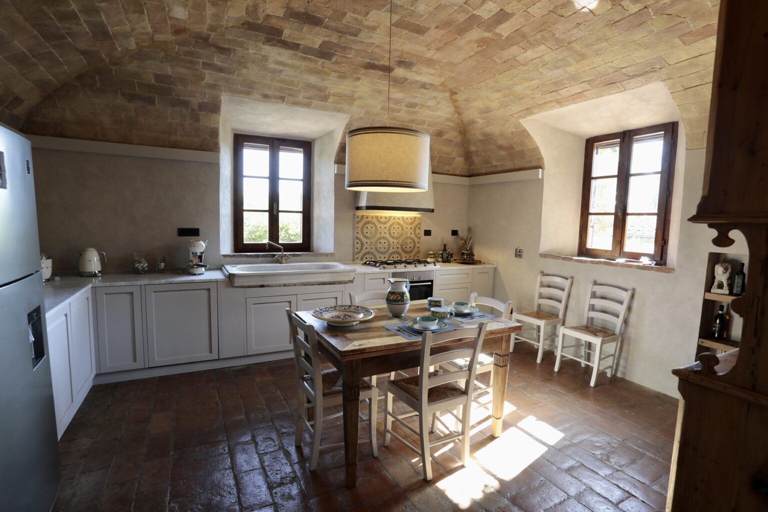 Bright and spacious rustic kitchen featuring a wooden dining table, white cabinetry, and large windows that allow natural light to fill the room. The ceiling has a unique arch design, and the floor is made of terracotta tiles. | Only on SopranoVillas