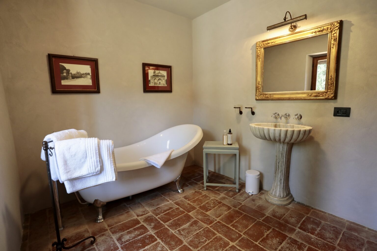 Elegant bathroom featuring a vintage freestanding bathtub, decorative wall art, a stylish mirror, and a classic pedestal sink on terracotta tile flooring. Perfect for showcasing luxurious home design. | Only on SopranoVillas