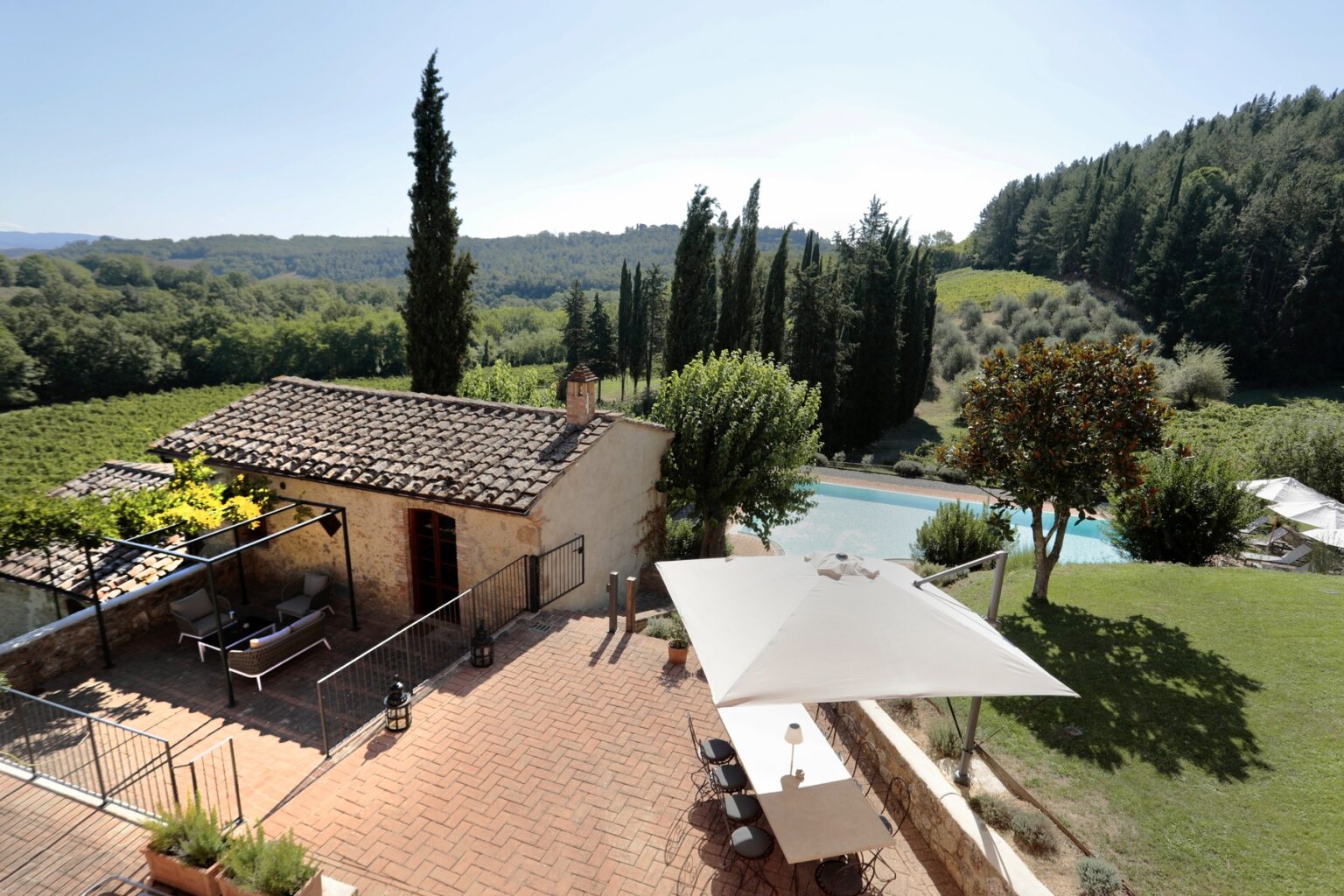 Scenic view of a rustic villa in Tuscany, featuring a stone building, lush greenery, and a sparkling swimming pool surrounded by cypress trees under a clear blue sky. Ideal for vacation rentals or countryside retreats. | Only on SopranoVillas