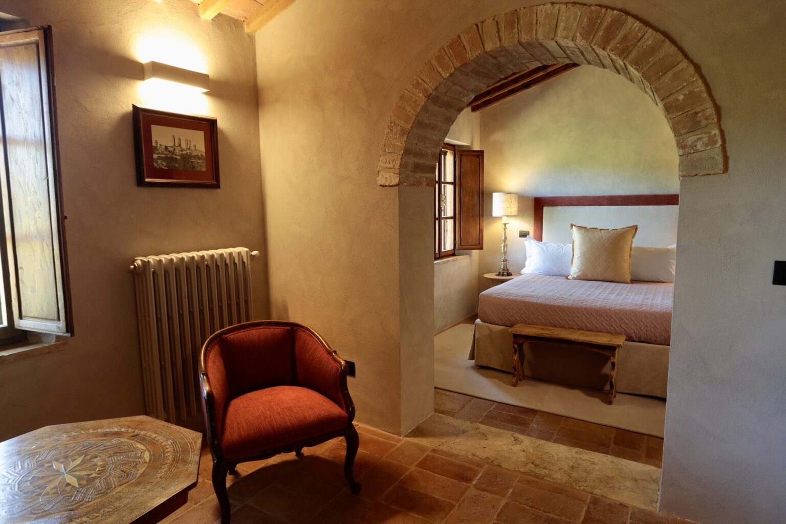 Cozy interior of a rustic bedroom featuring a comfortable bed, warm lighting, an elegant armchair, and an arched doorway leading to the sleeping area. The decor combines traditional elements with modern comfort, creating a welcoming atmosphere. | Only on SopranoVillas