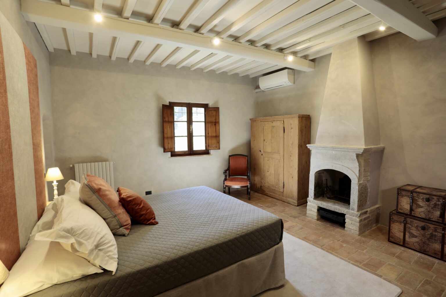 Cozy bedroom featuring a queen-sized bed with decorative pillows, wooden beams on the ceiling, a vintage wardrobe, and a traditional fireplace, complemented by natural light from a window. Ideal for relaxation and comfort. | Only on SopranoVillas