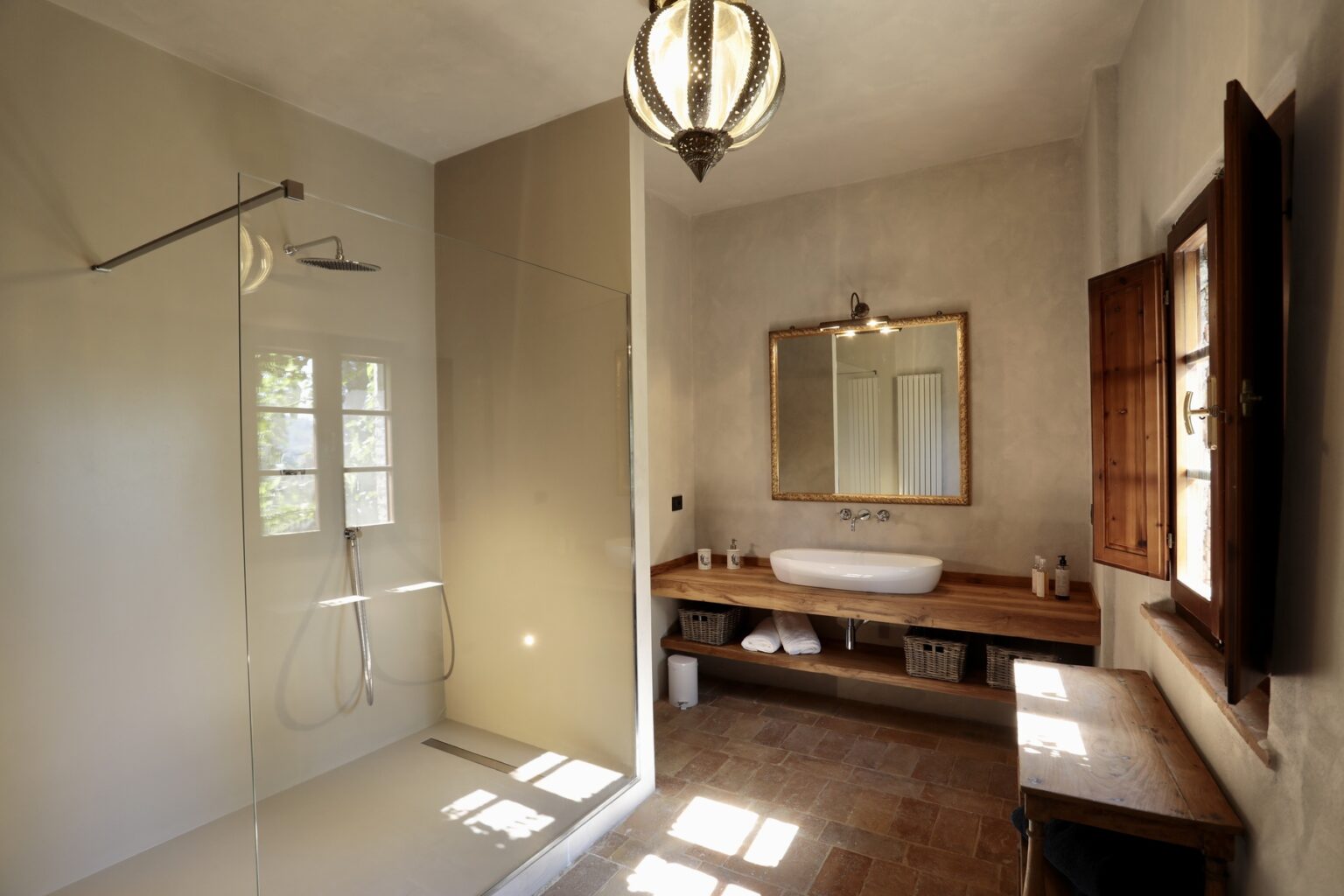 Modern bathroom interior featuring a spacious shower, a wooden vanity with a sink, a large mirror, and natural light from a window. The design combines minimalist elements with rustic touches, creating a serene atmosphere. | Only on SopranoVillas