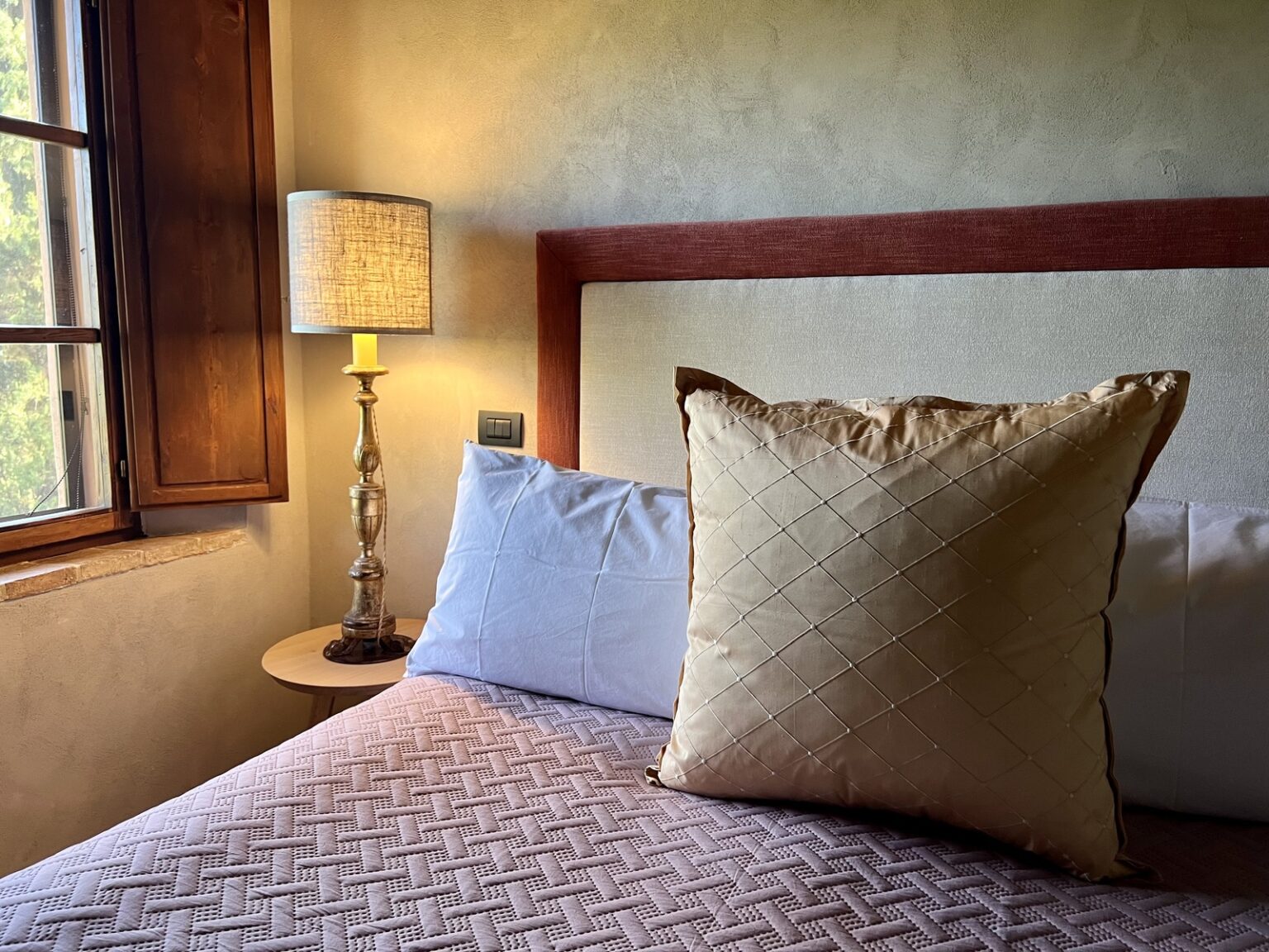 Cozy bedroom interior featuring a large decorative pillow on a neatly made bed, a bedside lamp, and a window allowing natural light to illuminate the space. | Only on SopranoVillas