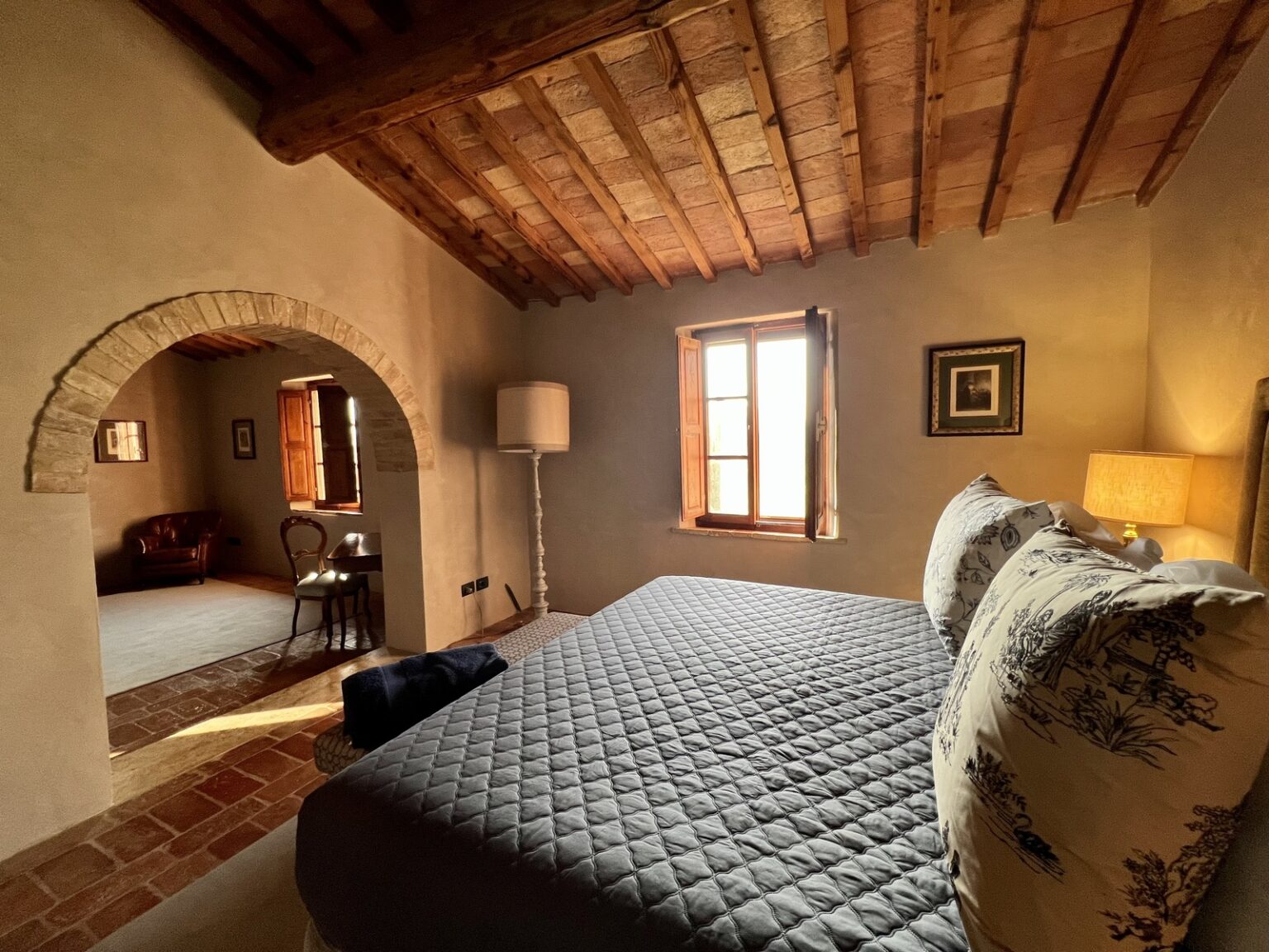 Cozy bedroom with a wooden ceiling, featuring a quilted bed, decorative pillows, and warm lighting from a bedside lamp. An arched doorway leads to a separate sitting area with a leather chair and wooden table, illuminated by natural light from a nearby window. Ideal for relaxation and comfort in a rustic setting. | Only on SopranoVillas