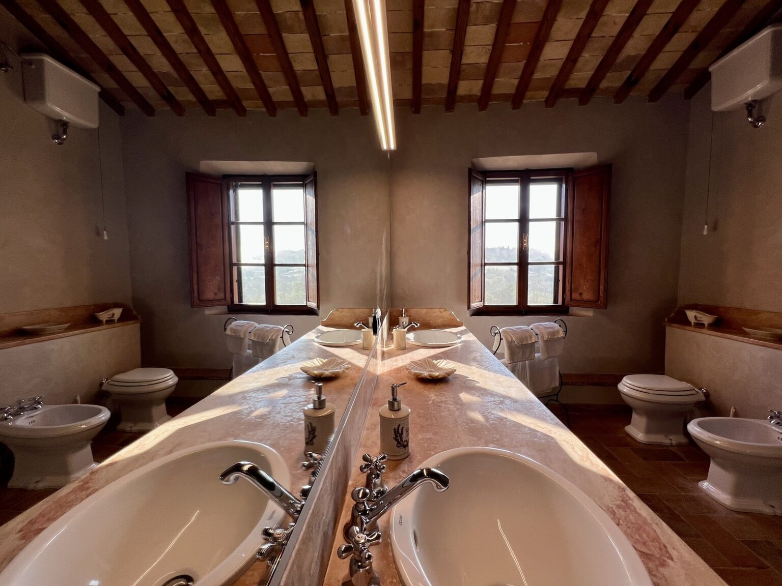 Modern bathroom interior featuring double sinks, wooden beams, and large windows with scenic views. The design combines elegance and functionality, showcasing a spacious layout with two toilets and stylish fixtures. | Only on SopranoVillas
