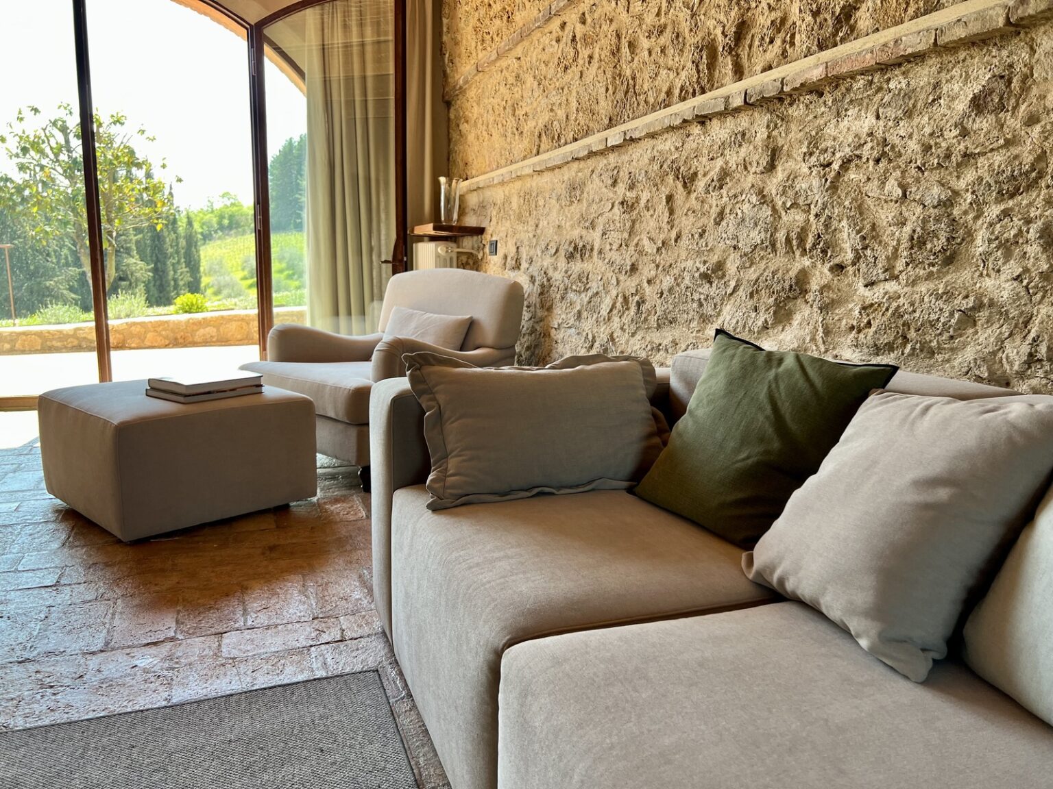 Cozy living room with a modern sofa and decorative pillows, featuring a stone wall and large windows that provide a view of the greenery outside. Natural light fills the space, enhancing the inviting atmosphere. | Only on SopranoVillas