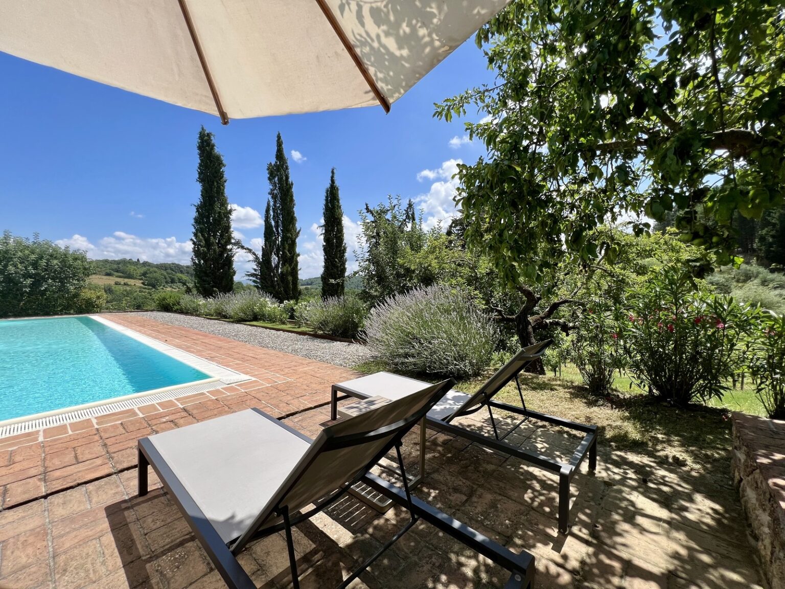 Lounge chairs beside a sparkling swimming pool surrounded by lush greenery and tall cypress trees under a blue sky, creating a tranquil outdoor retreat. | Only on SopranoVillas