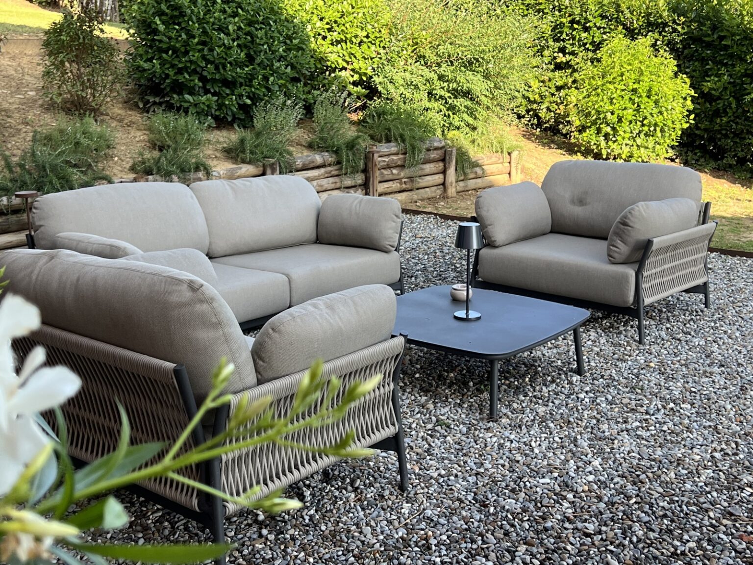 Modern outdoor seating arrangement featuring a gray fabric sofa set with plush cushions, accompanied by a sleek black coffee table, set in a landscaped garden with greenery. | Only on SopranoVillas