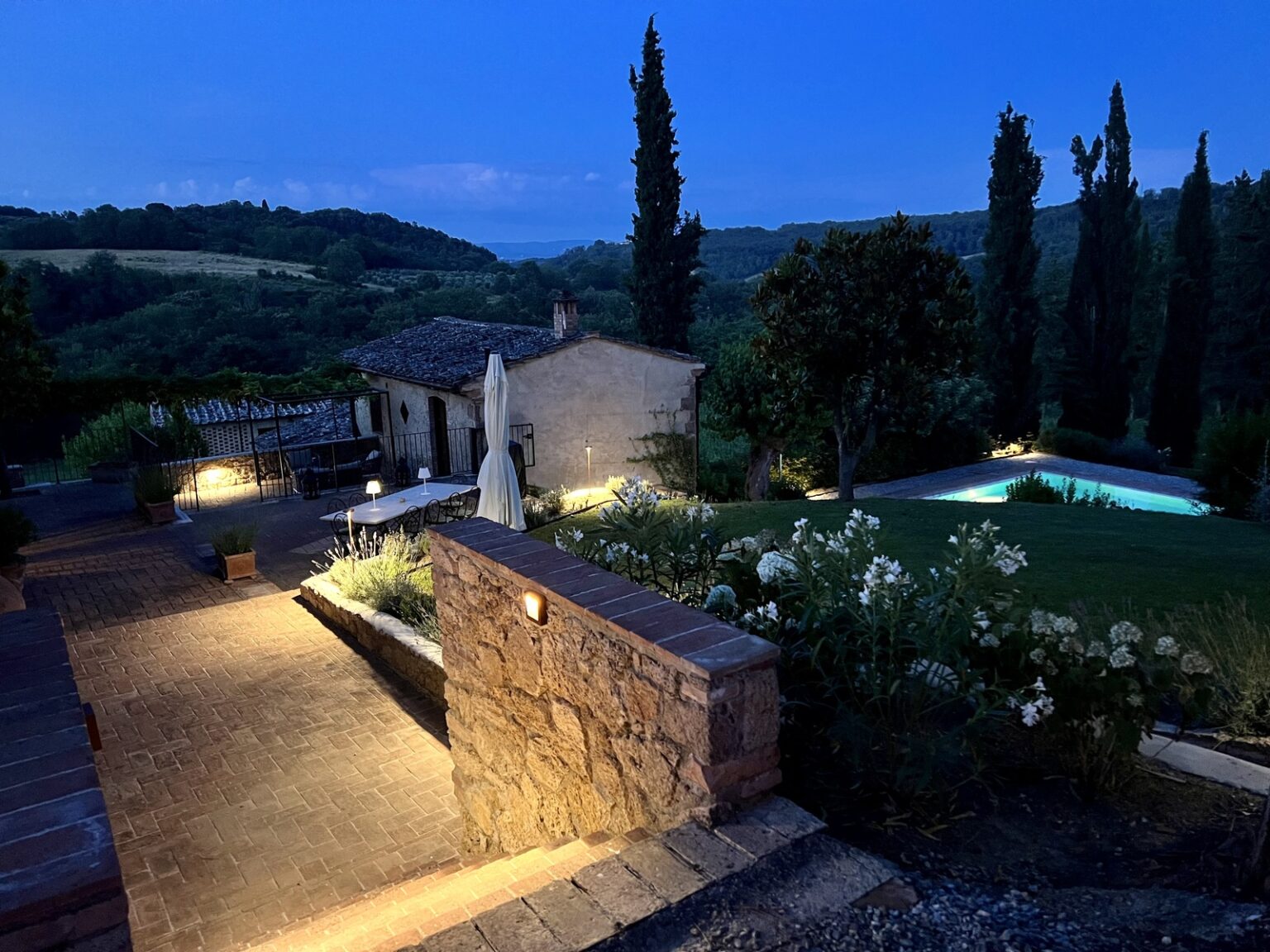 Scenic view of a tranquil countryside villa at dusk, featuring illuminated pathways, lush greenery, and a serene swimming pool surrounded by flowers and trees, set against a backdrop of rolling hills. | Only on SopranoVillas