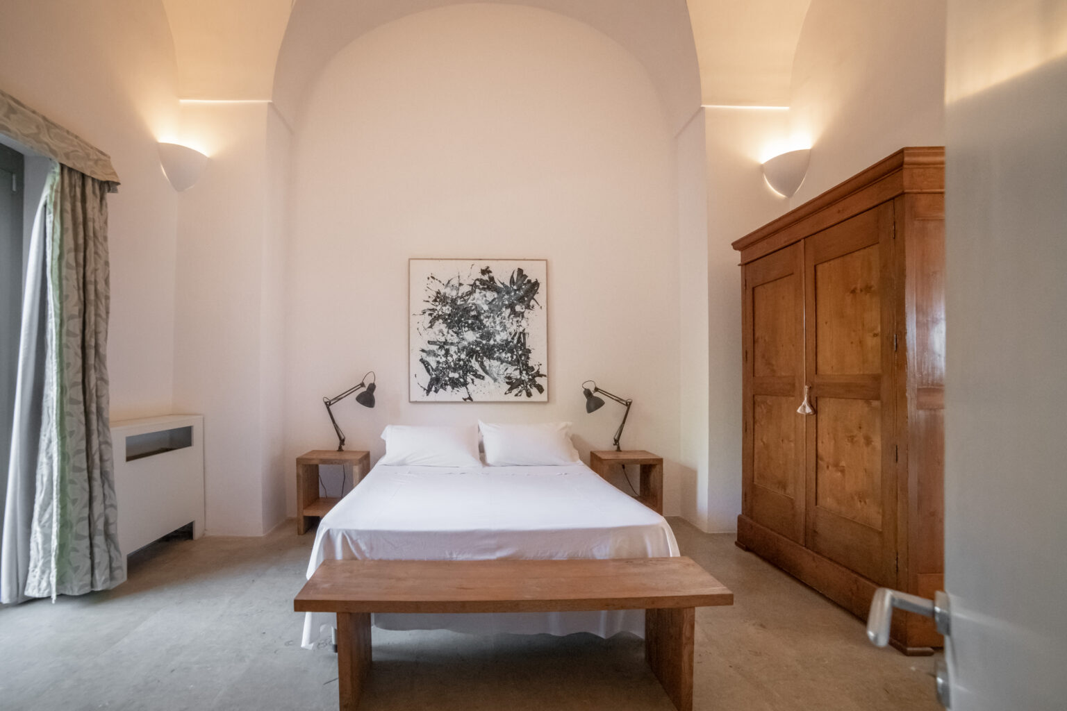 Modern bedroom interior featuring a double bed with white linens, two bedside tables with lamps, a wooden bench, and a large wooden wardrobe. The room has neutral walls and soft lighting, creating a serene atmosphere. | Only on SopranoVillas