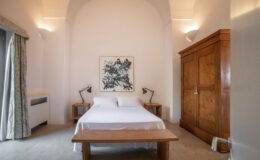 Modern bedroom interior featuring a double bed with white linens, two bedside tables with lamps, a wooden bench, and a large wooden wardrobe. The room has neutral walls and soft lighting, creating a serene atmosphere.