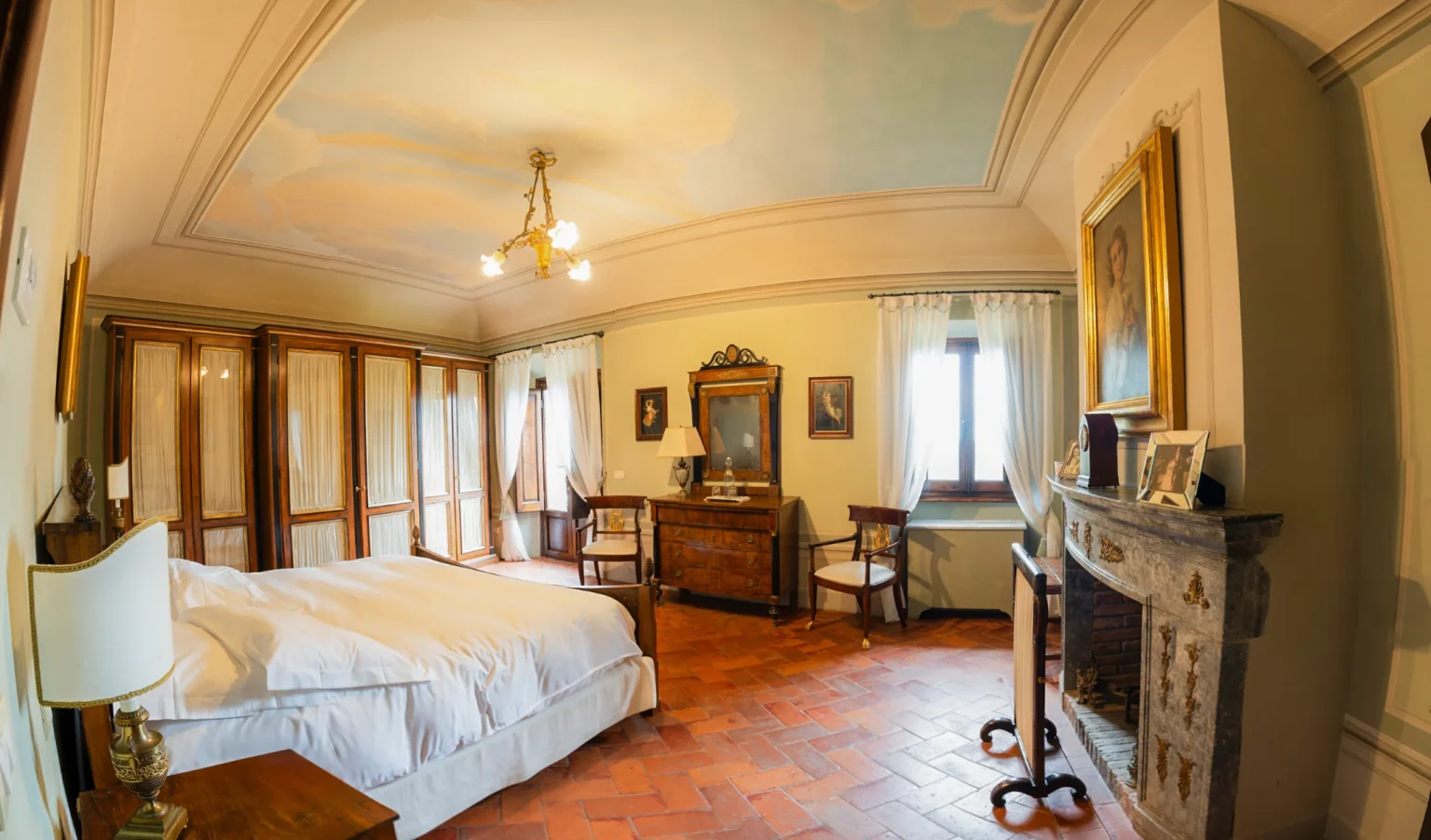 Elegant bedroom featuring a wooden bed with white linens, vintage furniture, and a decorative ceiling. The room includes a large wardrobe, a mirror, and two chairs, with natural light streaming through the windows adorned with sheer curtains. The terracotta floor adds warmth to the classic decor. | Only on SopranoVillas