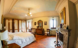 Elegant bedroom featuring a wooden bed with white linens, vintage furniture, and a decorative ceiling. The room includes a large wardrobe, a mirror, and two chairs, with natural light streaming through the windows adorned with sheer curtains. The terracotta floor adds warmth to the classic decor.