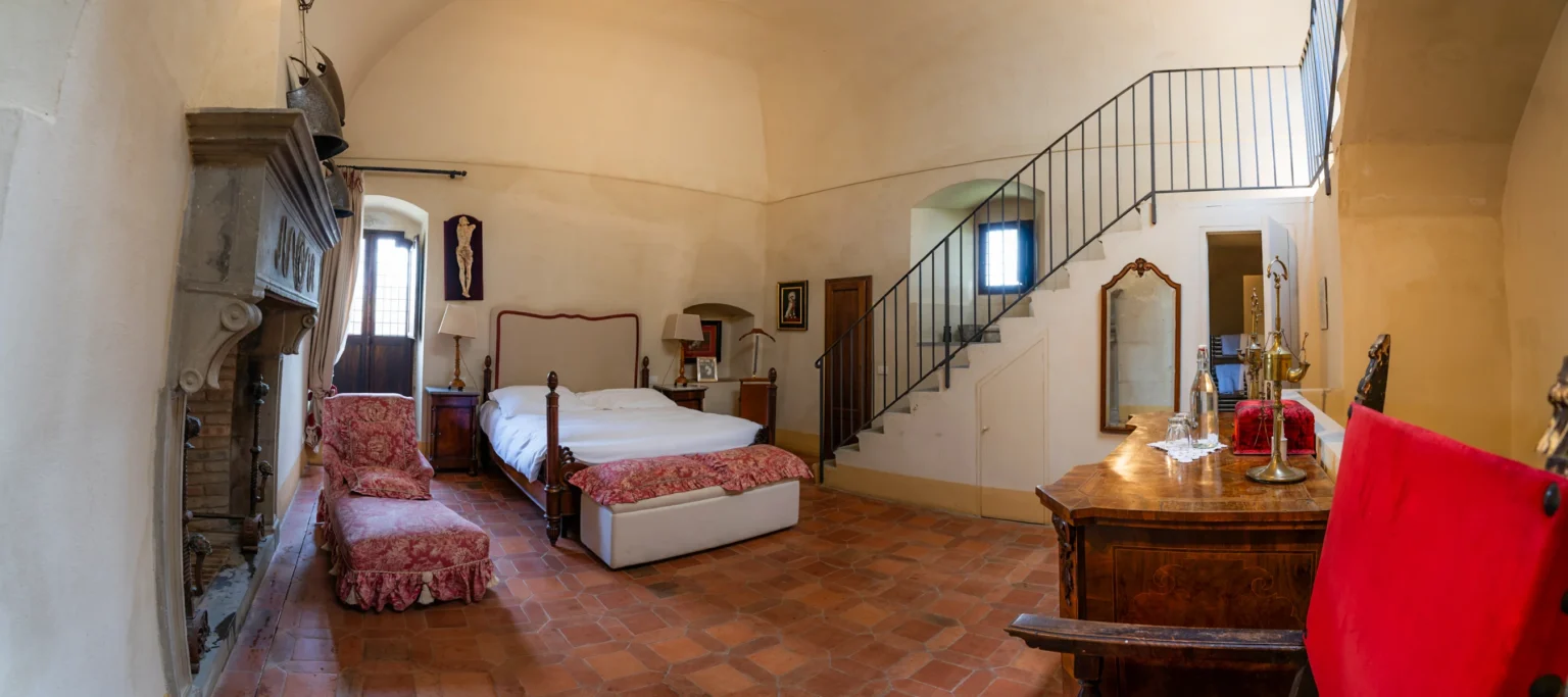 Spacious and elegantly designed bedroom featuring a wooden bed, vintage furniture, and a cozy seating area. The room has high ceilings, a decorative fireplace, and large windows allowing natural light. A staircase leads to an upper level, enhancing the room's charm and character. | Only on SopranoVillas