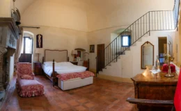 Spacious and elegantly designed bedroom featuring a wooden bed, vintage furniture, and a cozy seating area. The room has high ceilings, a decorative fireplace, and large windows allowing natural light. A staircase leads to an upper level, enhancing the room's charm and character.