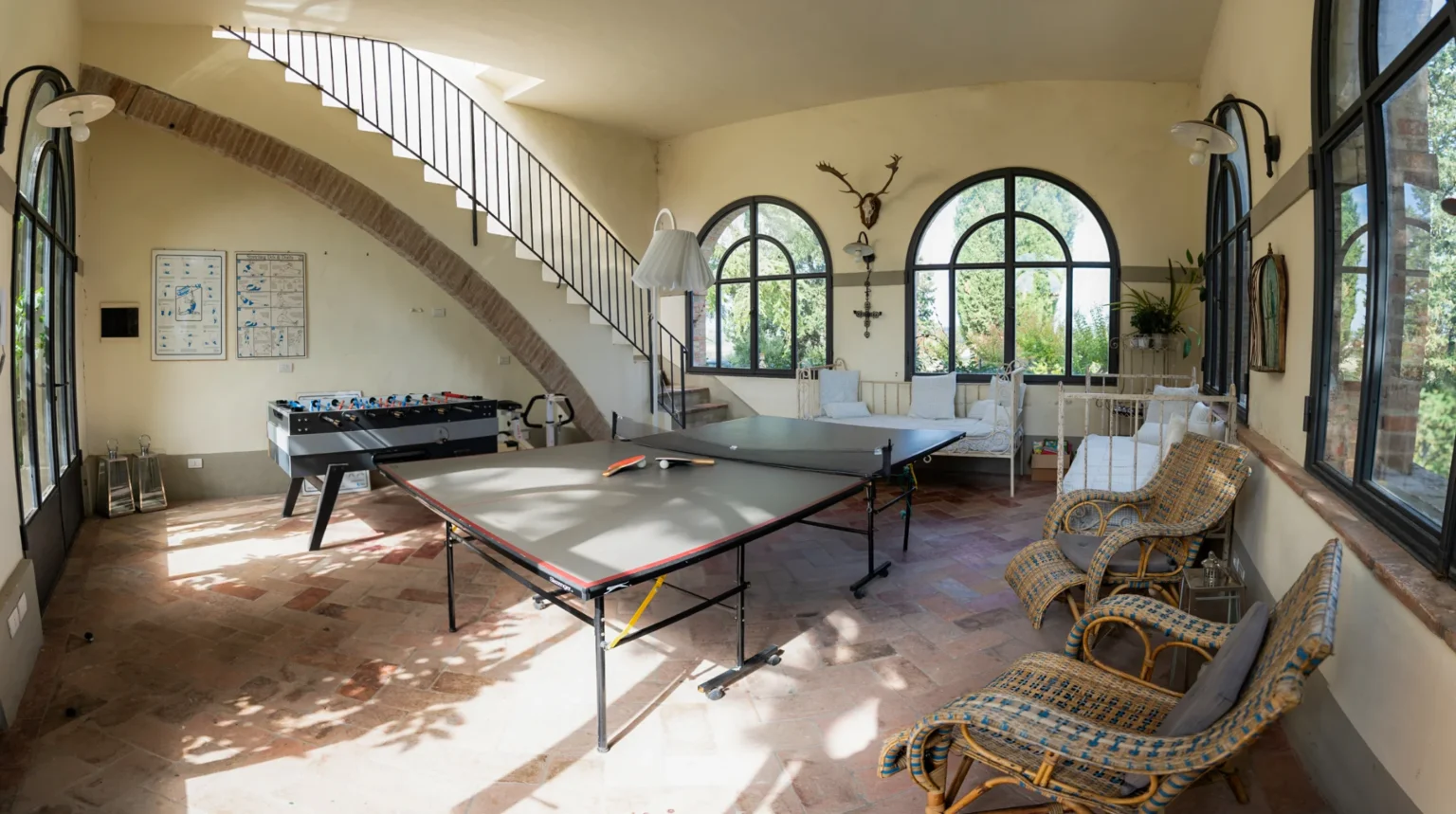 Spacious game room featuring two ping pong tables, a foosball table, and comfortable seating. Large windows provide natural light and views of the surrounding greenery, enhancing the inviting atmosphere. | Only on SopranoVillas