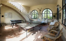 Spacious game room featuring two ping pong tables, a foosball table, and comfortable seating. Large windows provide natural light and views of the surrounding greenery, enhancing the inviting atmosphere.
