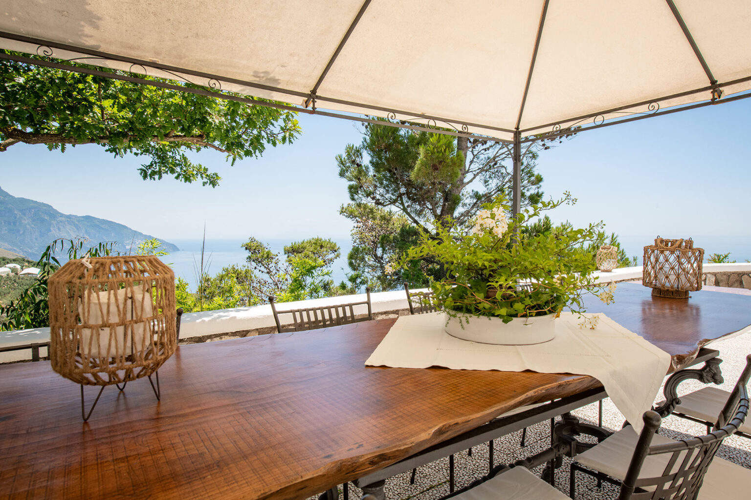 Outdoor dining area with a wooden table, surrounded by chairs, under a canopy. Lush greenery and a stunning ocean view create a serene atmosphere, perfect for al fresco dining. Decorative lanterns enhance the inviting ambiance. | Only on SopranoVillas