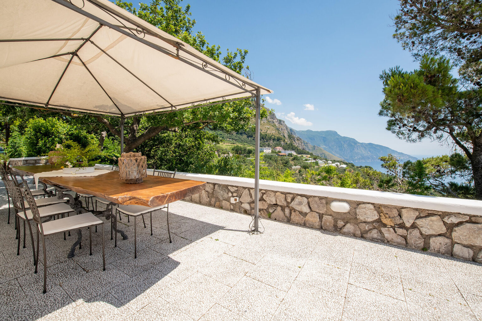 Outdoor dining area with a large wooden table and metal chairs under a shaded canopy, overlooking a scenic view of mountains and coastline. Perfect for al fresco dining and gatherings. | Only on SopranoVillas