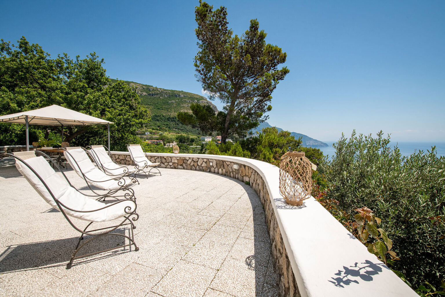 Outdoor terrace with sun loungers overlooking a scenic coastal view, surrounded by lush greenery and a shaded area with an umbrella. Ideal for relaxation and enjoying the sun. | Only on SopranoVillas
