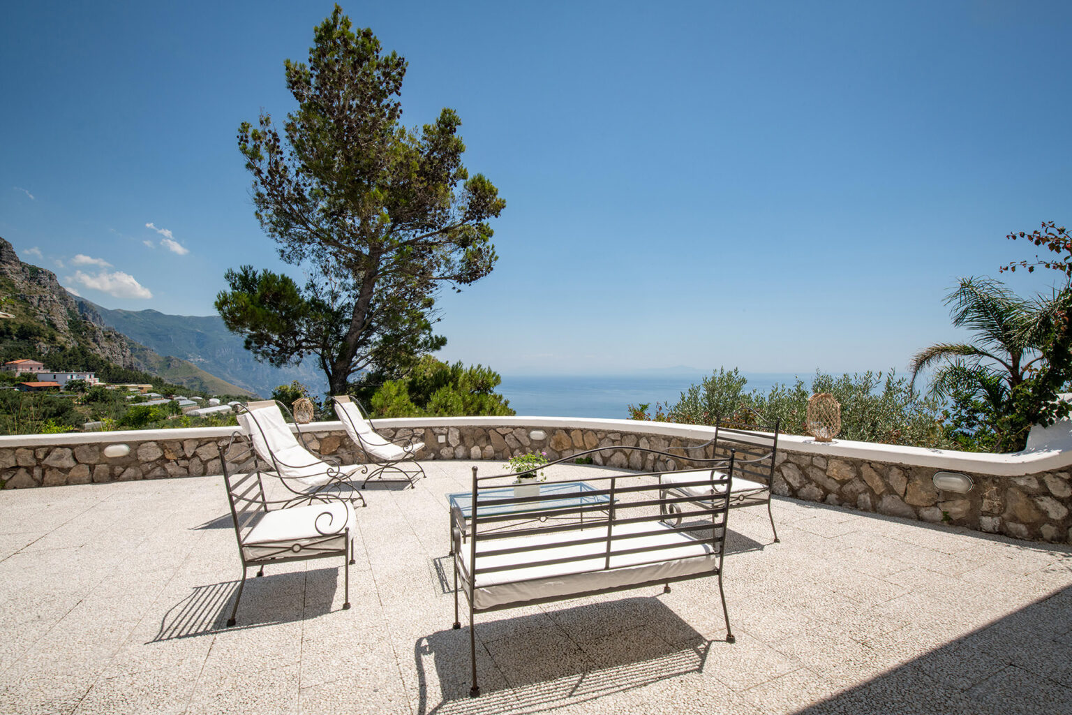 Outdoor terrace with metal furniture overlooking the ocean and mountains under a clear blue sky. Perfect for relaxation and enjoying scenic views. | Only on SopranoVillas