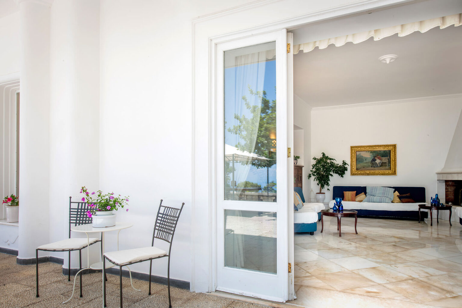 Bright and airy interior of a modern villa showcasing a living room with a blue sofa and artwork, viewed from an open door leading to a small outdoor seating area with two chairs and potted flowers. | Only on SopranoVillas