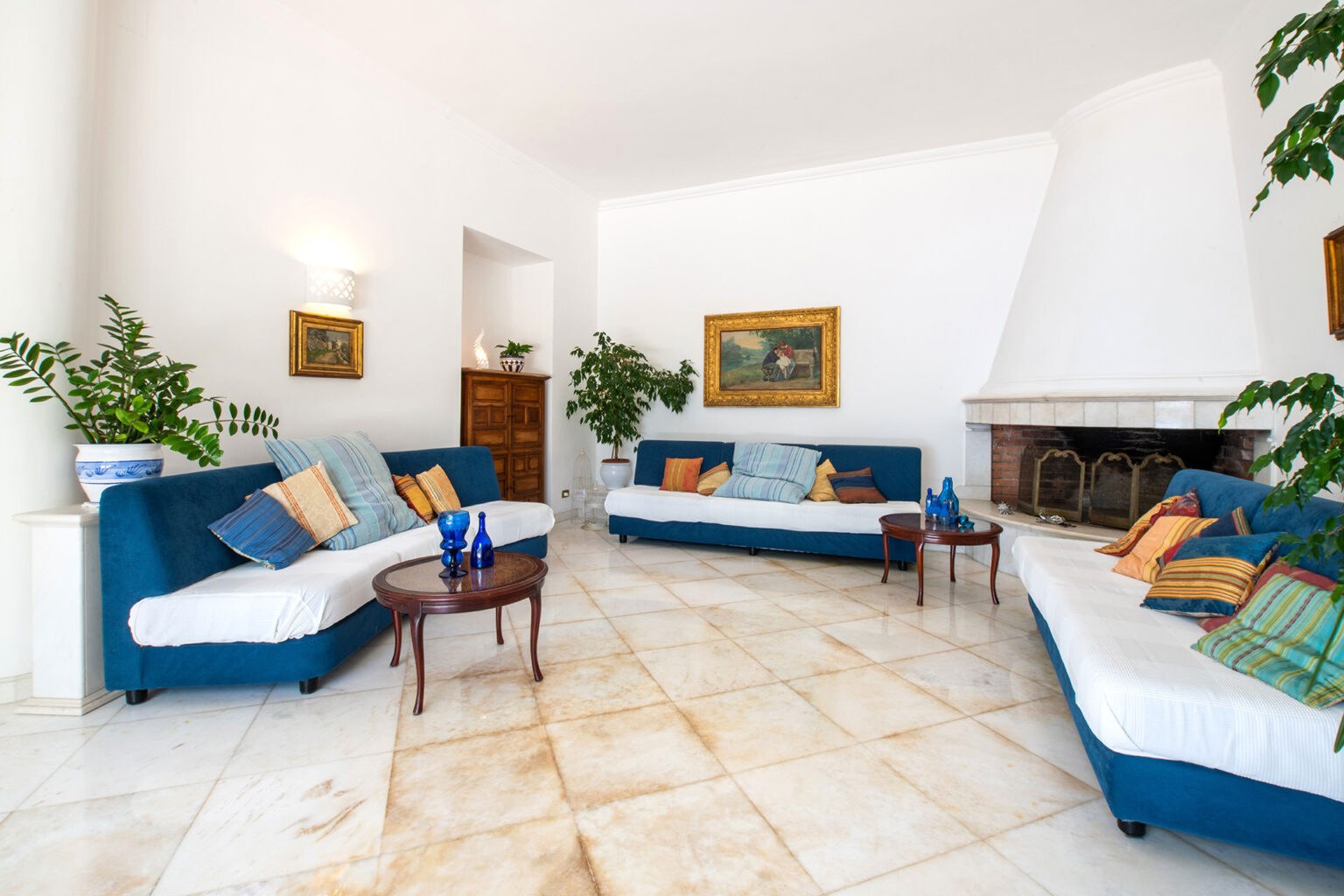 Bright and spacious living room featuring blue sofas adorned with colorful cushions, a decorative coffee table, and indoor plants, complemented by a cozy fireplace and artwork on the walls. | Only on SopranoVillas