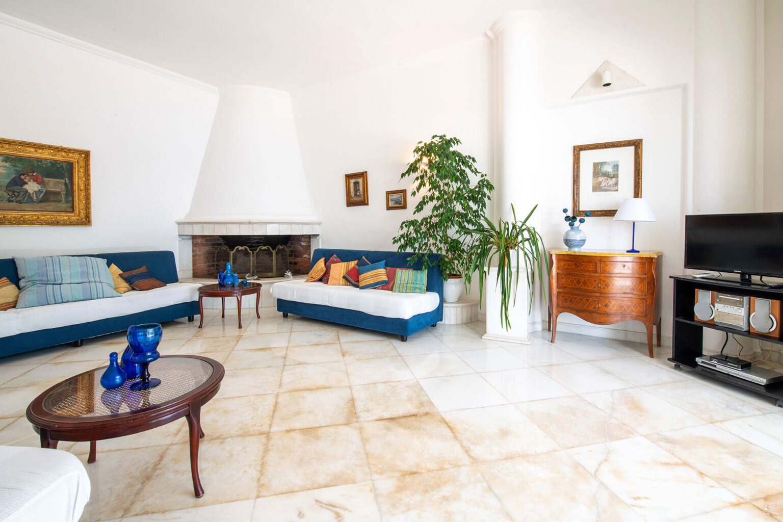 Bright and airy living room featuring blue sofas, a coffee table, and a decorative plant, with a fireplace and television in the background, showcasing a modern yet cozy design. | Only on SopranoVillas