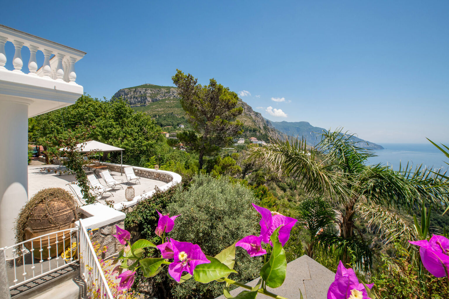 Scenic view from a villa terrace overlooking lush green hills and the ocean, featuring vibrant pink bougainvillea flowers, sun loungers, and a clear blue sky. Ideal for vacation rentals in a coastal destination. | Only on SopranoVillas
