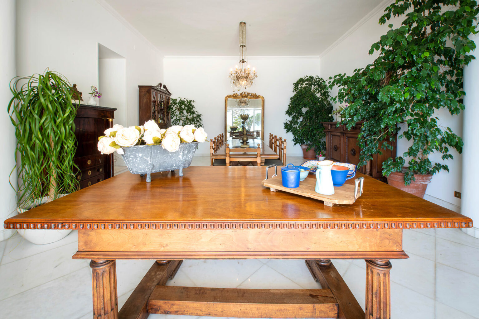 Spacious dining room featuring a wooden table adorned with white roses, blue cups, and a serving tray, surrounded by lush green plants and elegant furniture, creating a bright and inviting atmosphere. | Only on SopranoVillas