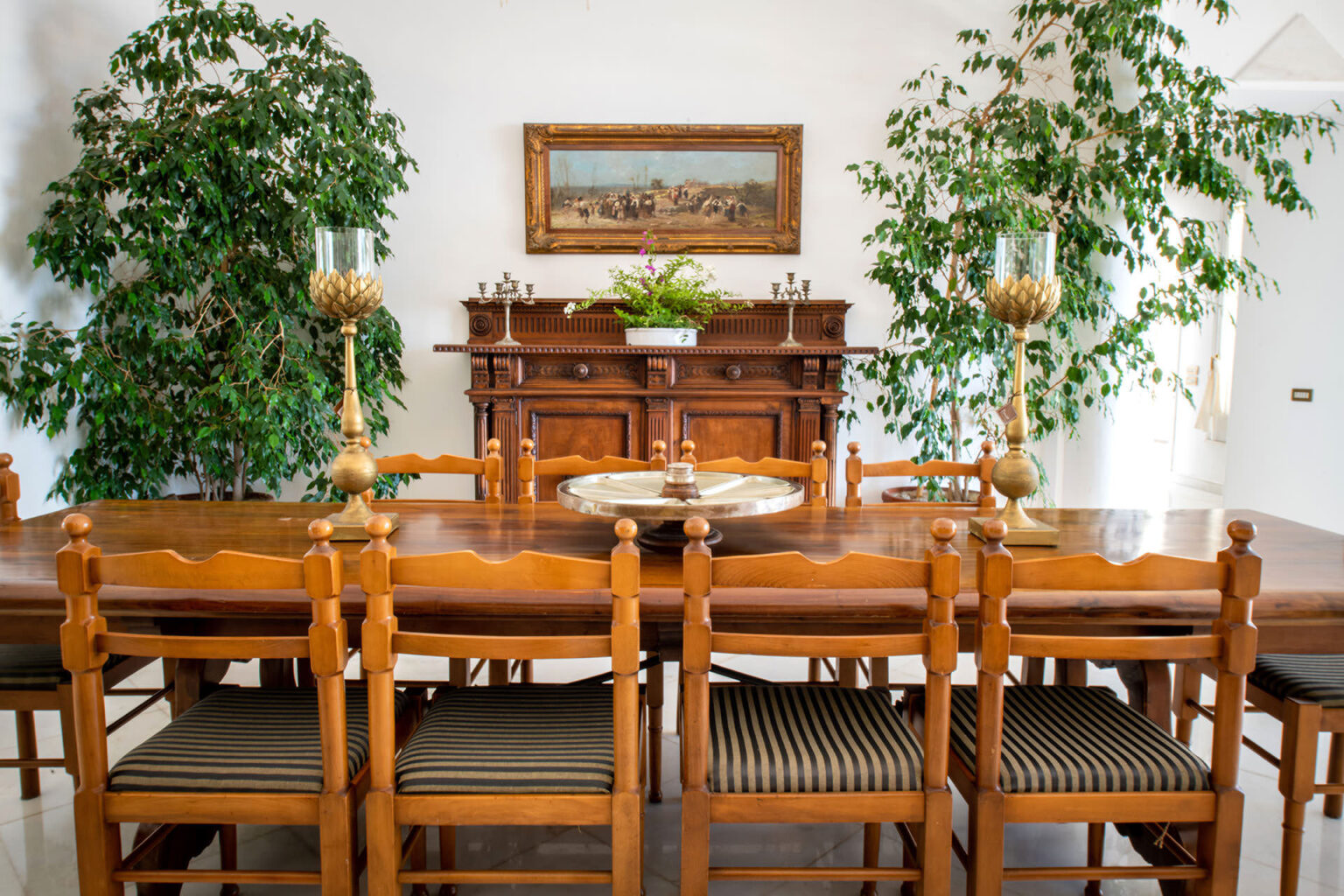 Spacious dining room featuring a wooden table with striped chairs, decorative gold candle holders, and lush greenery. A vintage sideboard with intricate detailing and a framed landscape painting adorn the wall, creating an elegant and inviting atmosphere. | Only on SopranoVillas