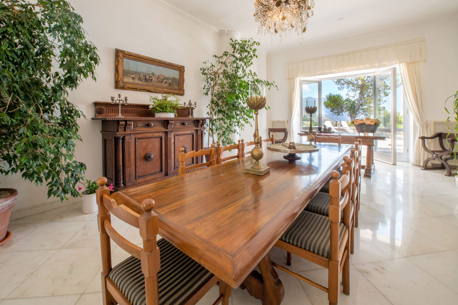 Elegant dining room featuring a large wooden table surrounded by striped chairs, a vintage sideboard, and decorative plants. A chandelier hangs from the ceiling, and large windows provide ample natural light and a view of the outdoors. | Only on SopranoVillas