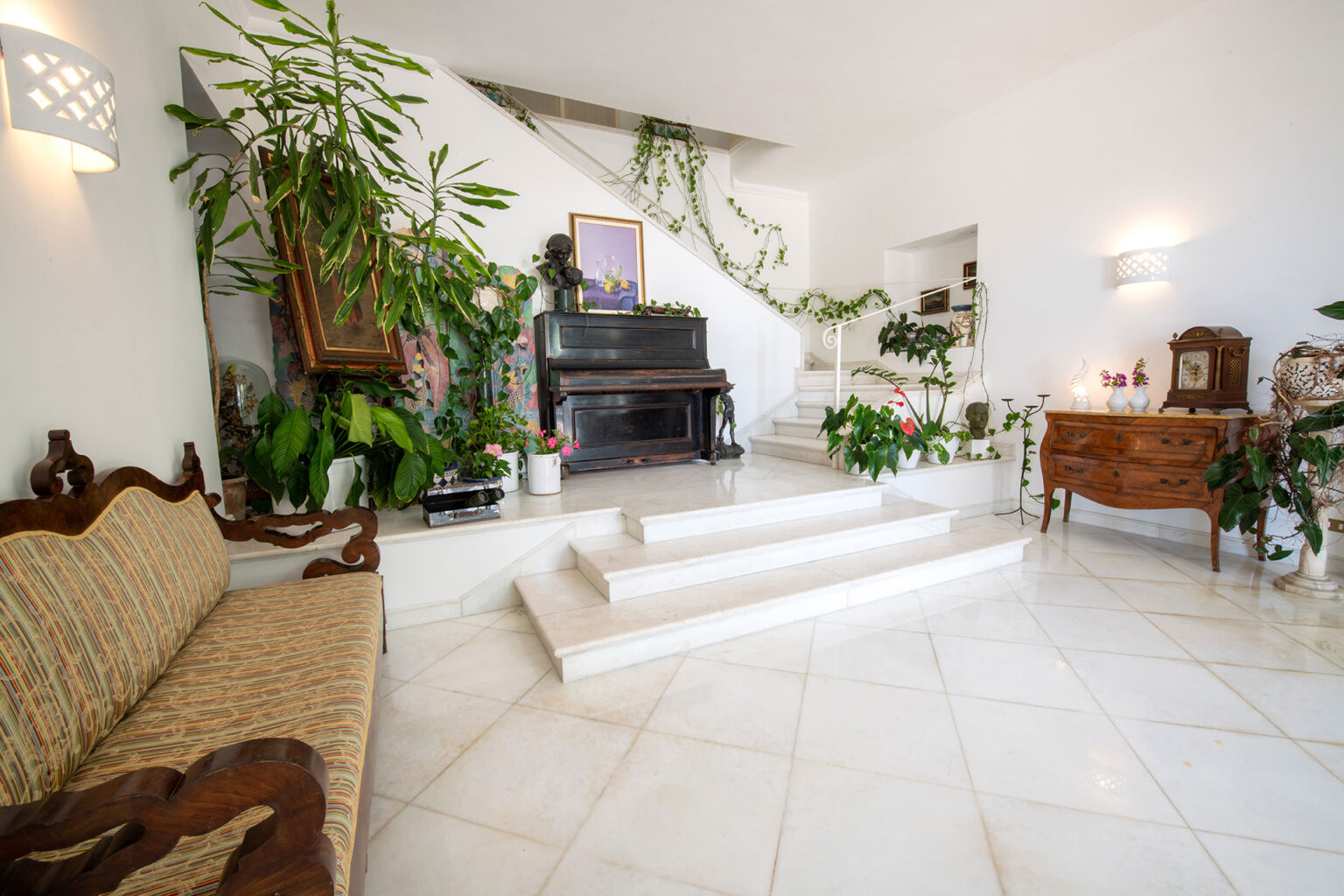 Bright and airy living room featuring a grand piano, elegant wooden furniture, and lush indoor plants. The space includes a staircase adorned with greenery, a vintage cabinet, and decorative art pieces, all set against a clean white backdrop. Ideal for showcasing interior design and home decor inspiration. | Only on SopranoVillas