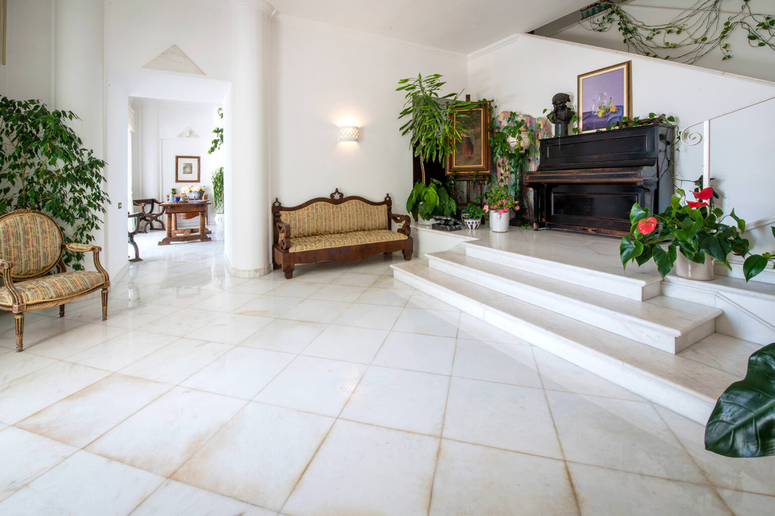 Bright and spacious living room featuring marble flooring, stylish vintage seating, and lush indoor plants. A classic piano and decorative artwork enhance the elegant ambiance, creating a welcoming atmosphere. Natural light floods the space through open doorways. | Only on SopranoVillas