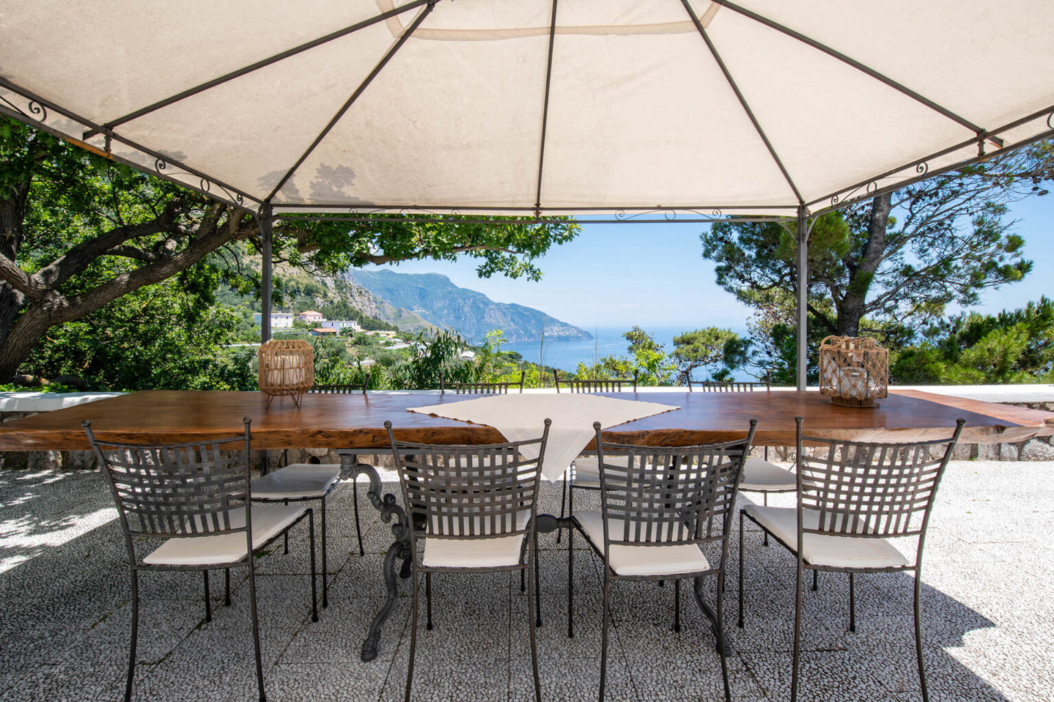 Outdoor dining area with a wooden table and metal chairs under a canopy, overlooking a scenic view of mountains and the sea. Ideal for al fresco dining in a tranquil setting. | Only on SopranoVillas