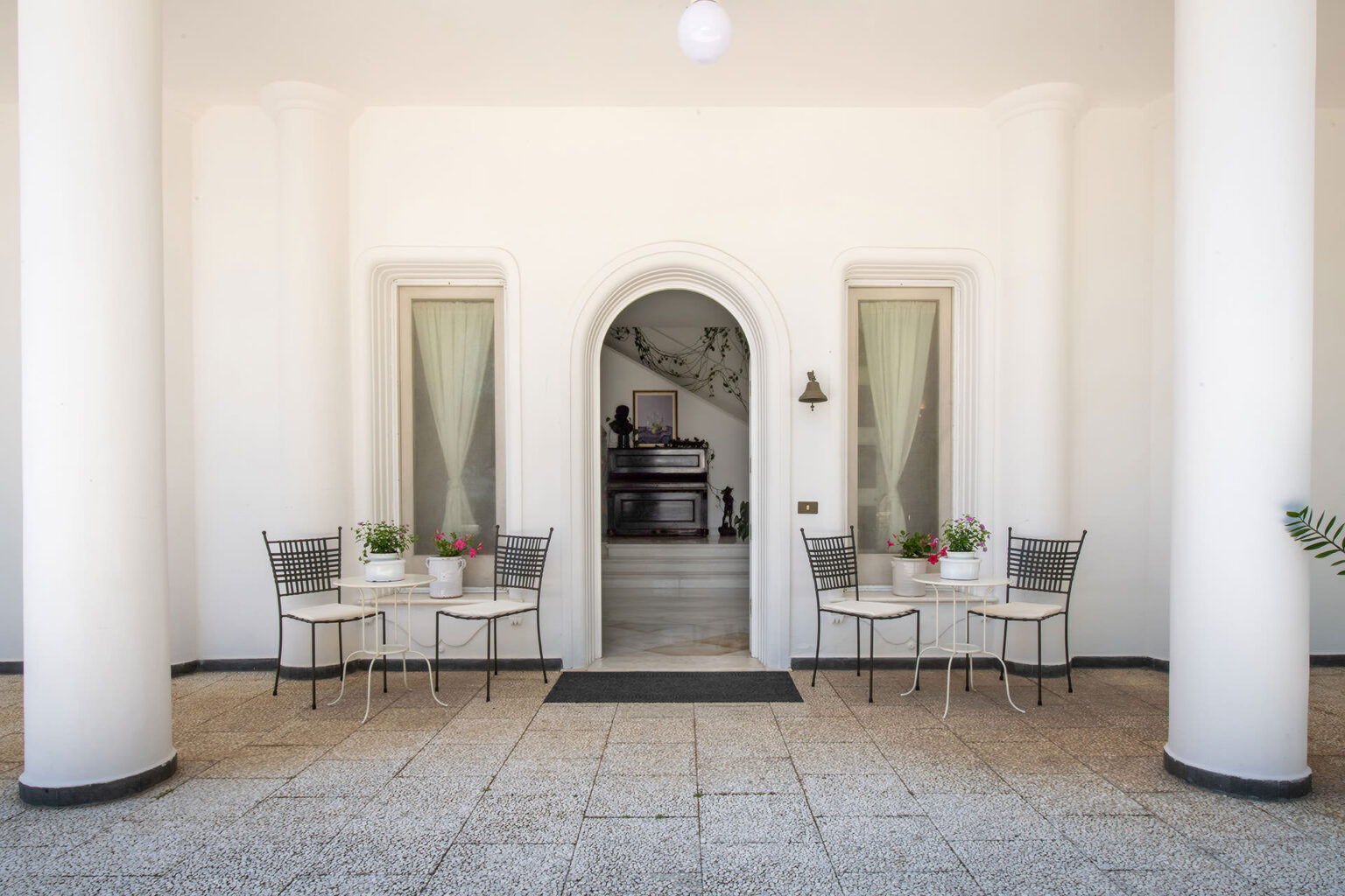 Charming outdoor café entrance with two tables and chairs, surrounded by white columns and potted plants, inviting guests to relax in a serene atmosphere. | Only on SopranoVillas
