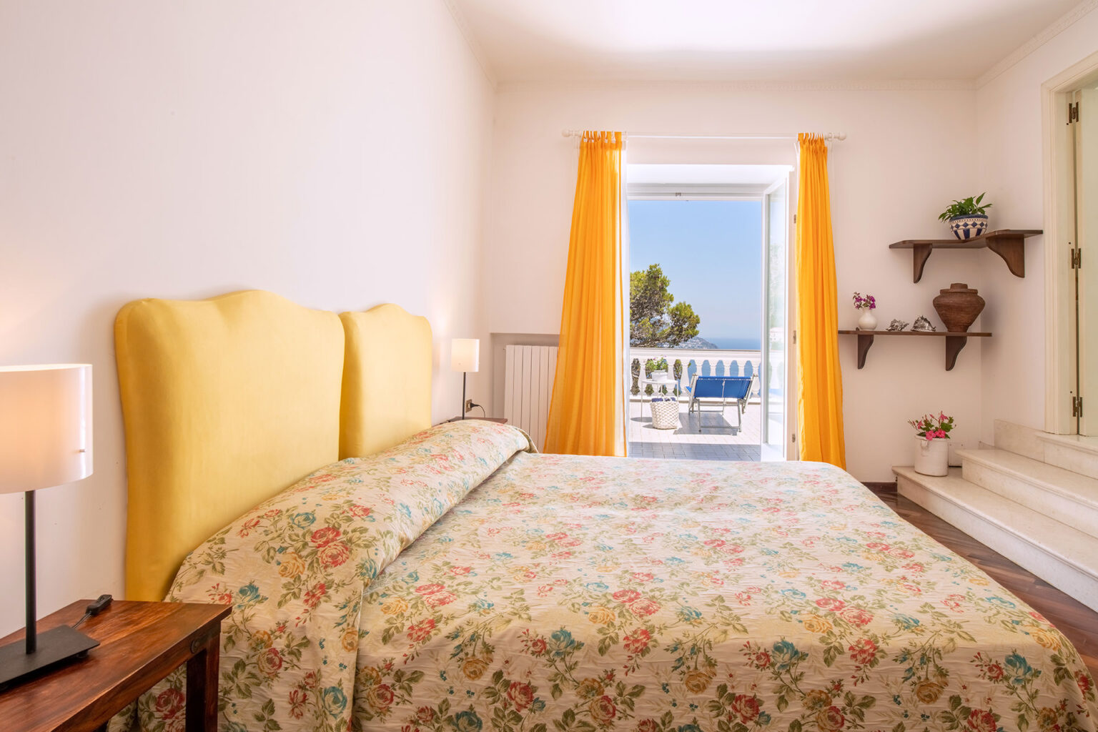 Bright and airy bedroom featuring a yellow upholstered headboard, floral bedding, and bedside lamps. The room opens to a scenic terrace with blue lounge chairs and views of the sea, complemented by vibrant yellow curtains and decorative shelves. Ideal for a relaxing getaway. | Only on SopranoVillas