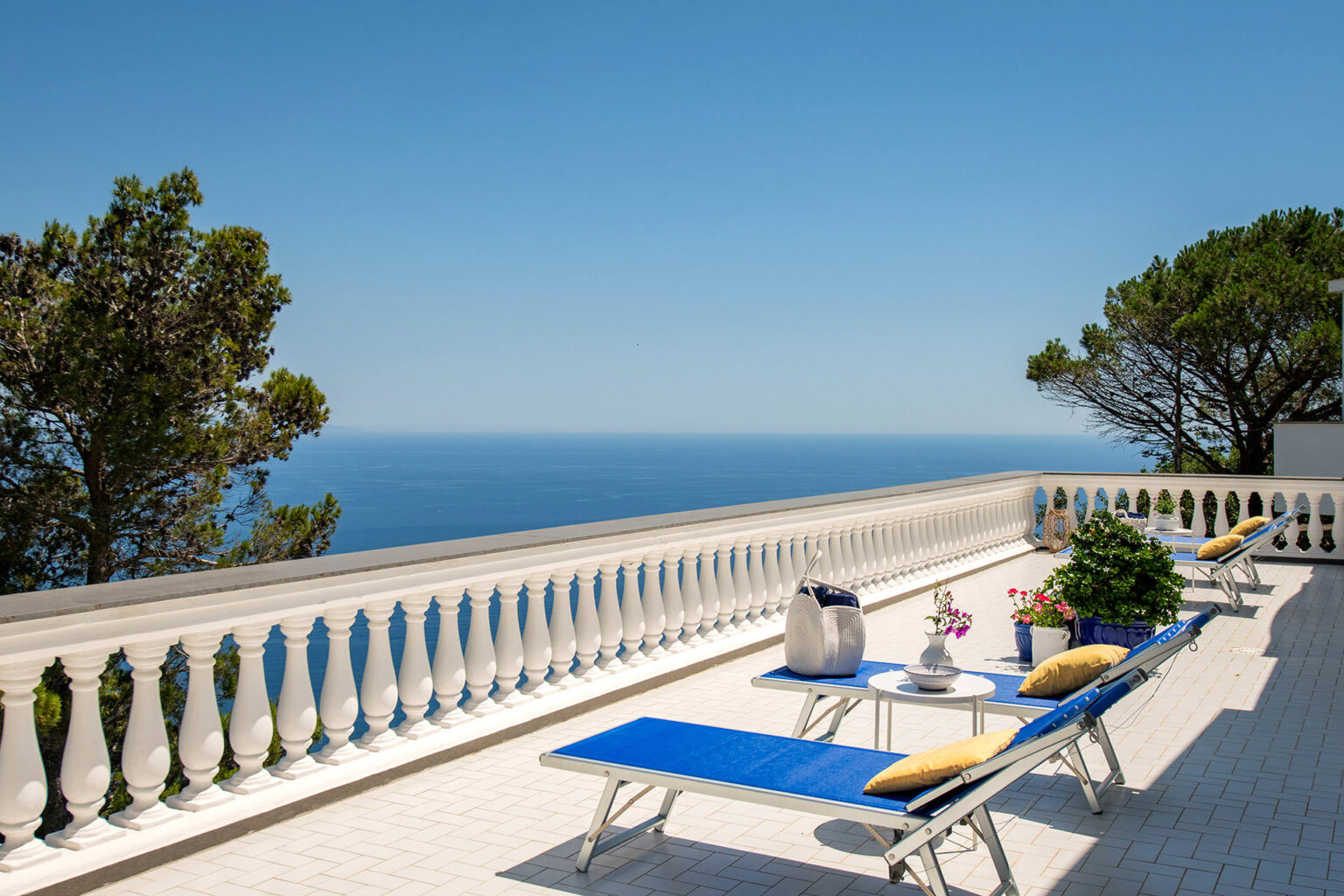 Luxury terrace with sun loungers overlooking the ocean, featuring a white railing and potted plants, ideal for relaxation and enjoying scenic coastal views. | Only on SopranoVillas