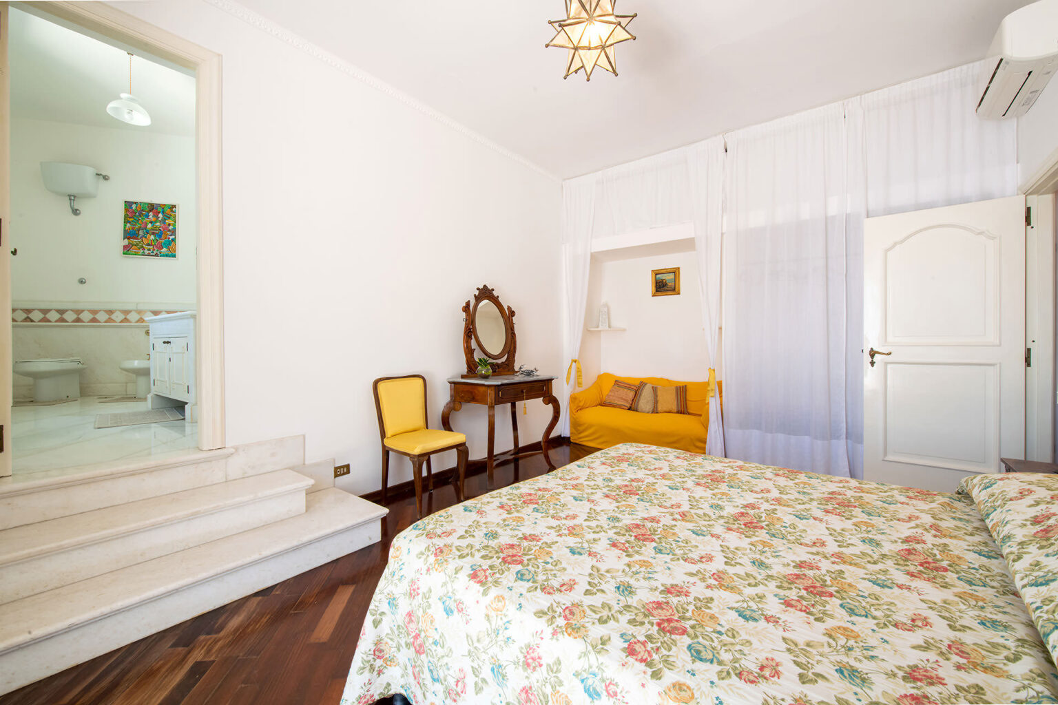 Bright and airy bedroom featuring a floral bedspread, a vintage mirror, and a cozy yellow sofa. A doorway leads to a modern bathroom with white fixtures. Natural light filters through sheer curtains, enhancing the inviting ambiance. | Only on SopranoVillas