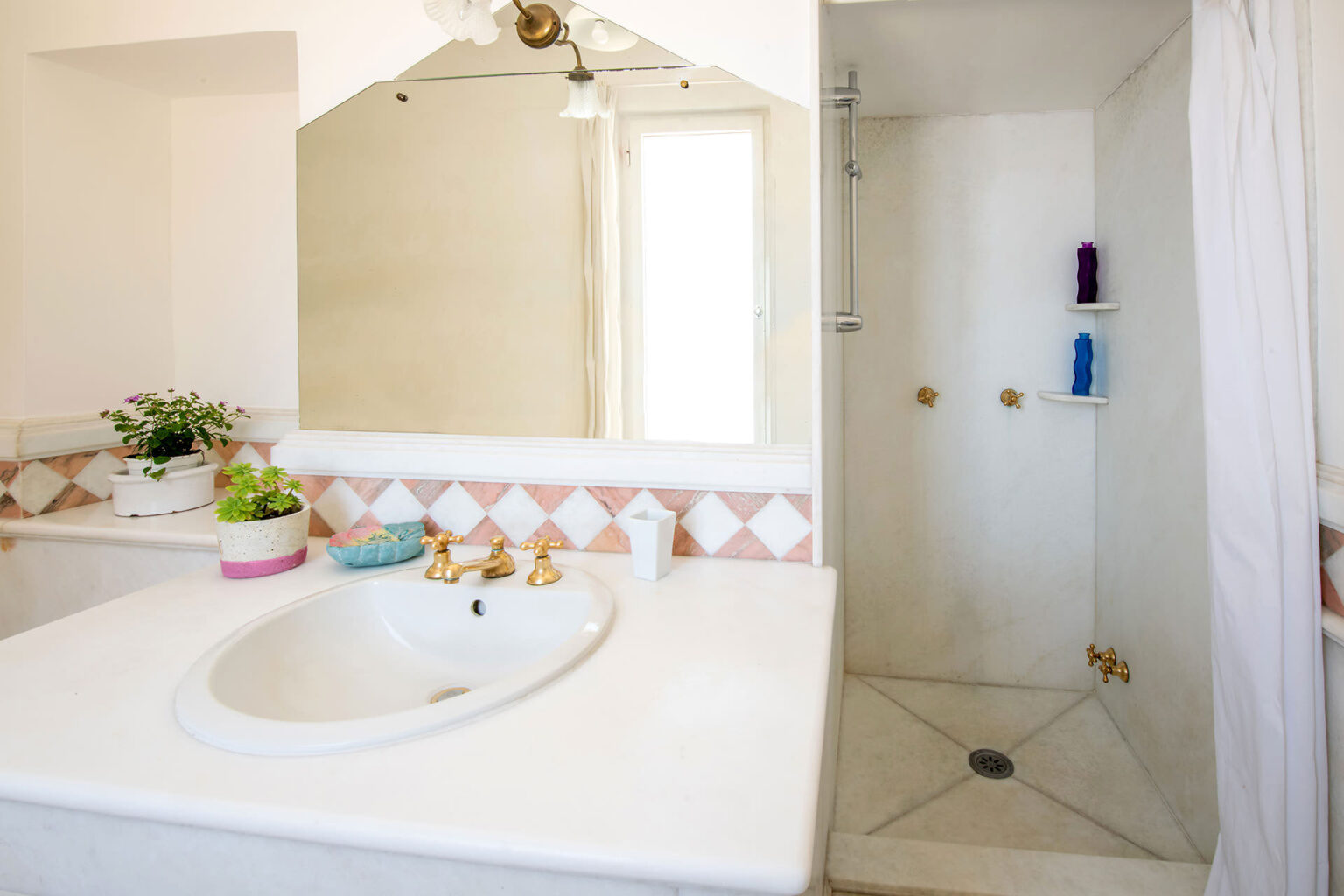 Bright and modern bathroom featuring a white sink with gold fixtures, a large mirror, and a walk-in shower. Decorative elements include potted plants and colorful toiletries on display, complemented by light-colored tiles and a serene atmosphere. | Only on SopranoVillas
