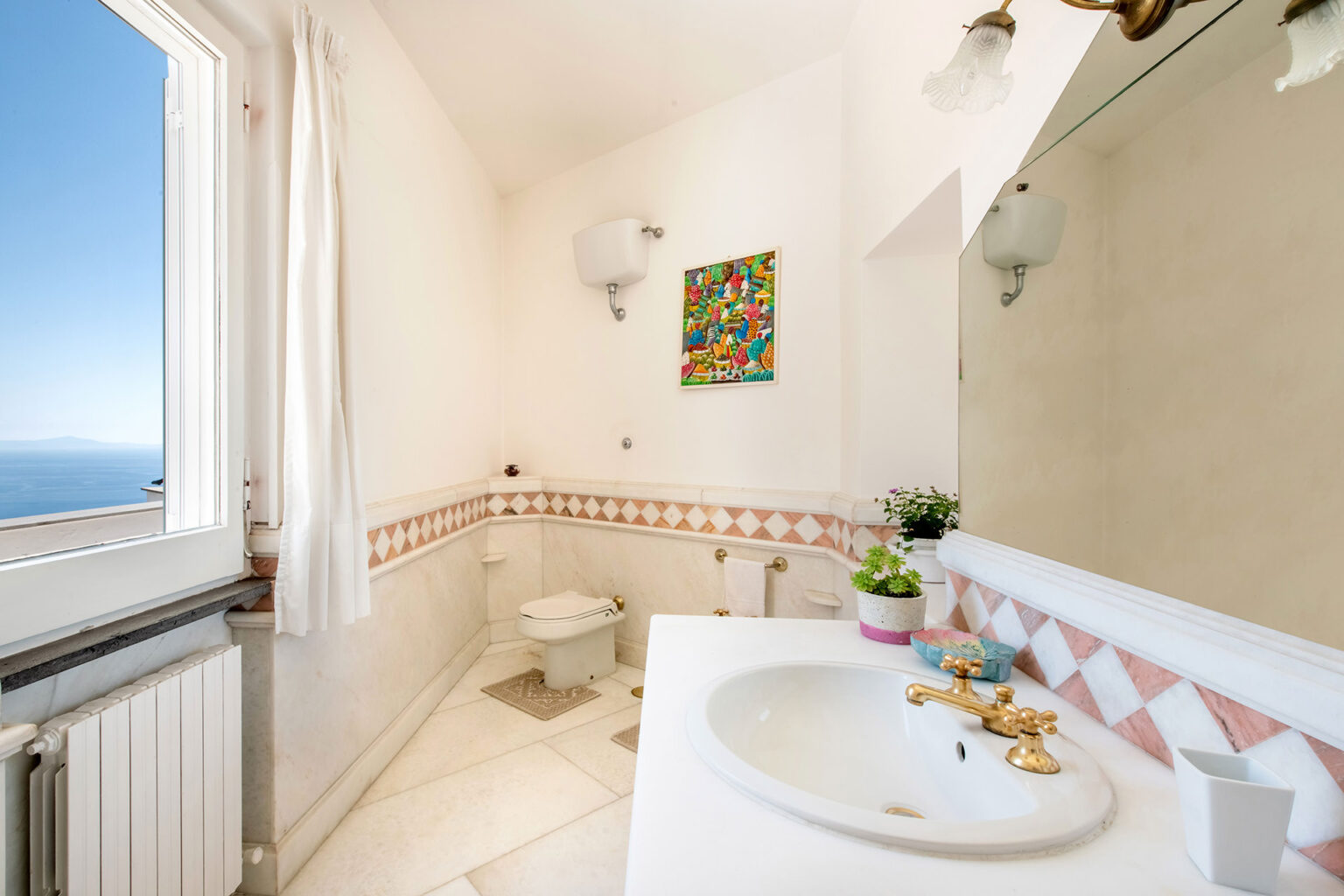 Bright and airy bathroom with a view, featuring a white sink, decorative wall art, and a tiled border. Natural light floods the space through large windows, creating a serene atmosphere. | Only on SopranoVillas