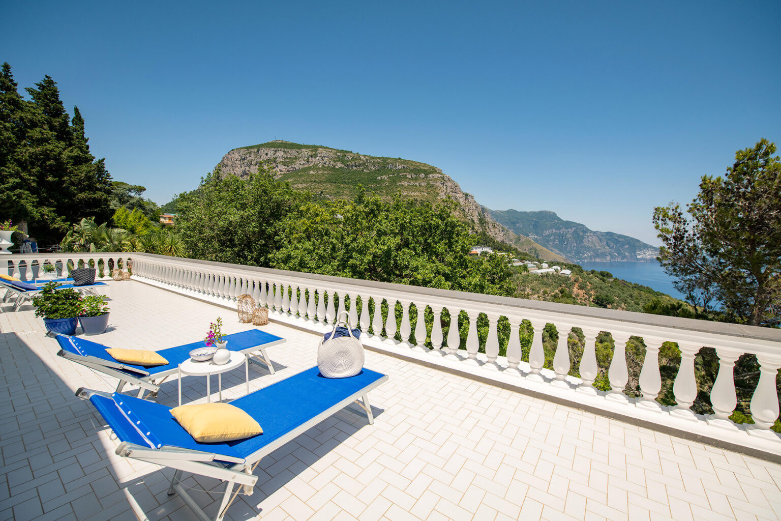 Scenic terrace with blue lounge chairs, overlooking a lush green mountain and coastline under a clear blue sky. Ideal for relaxation and enjoying outdoor views. | Only on SopranoVillas