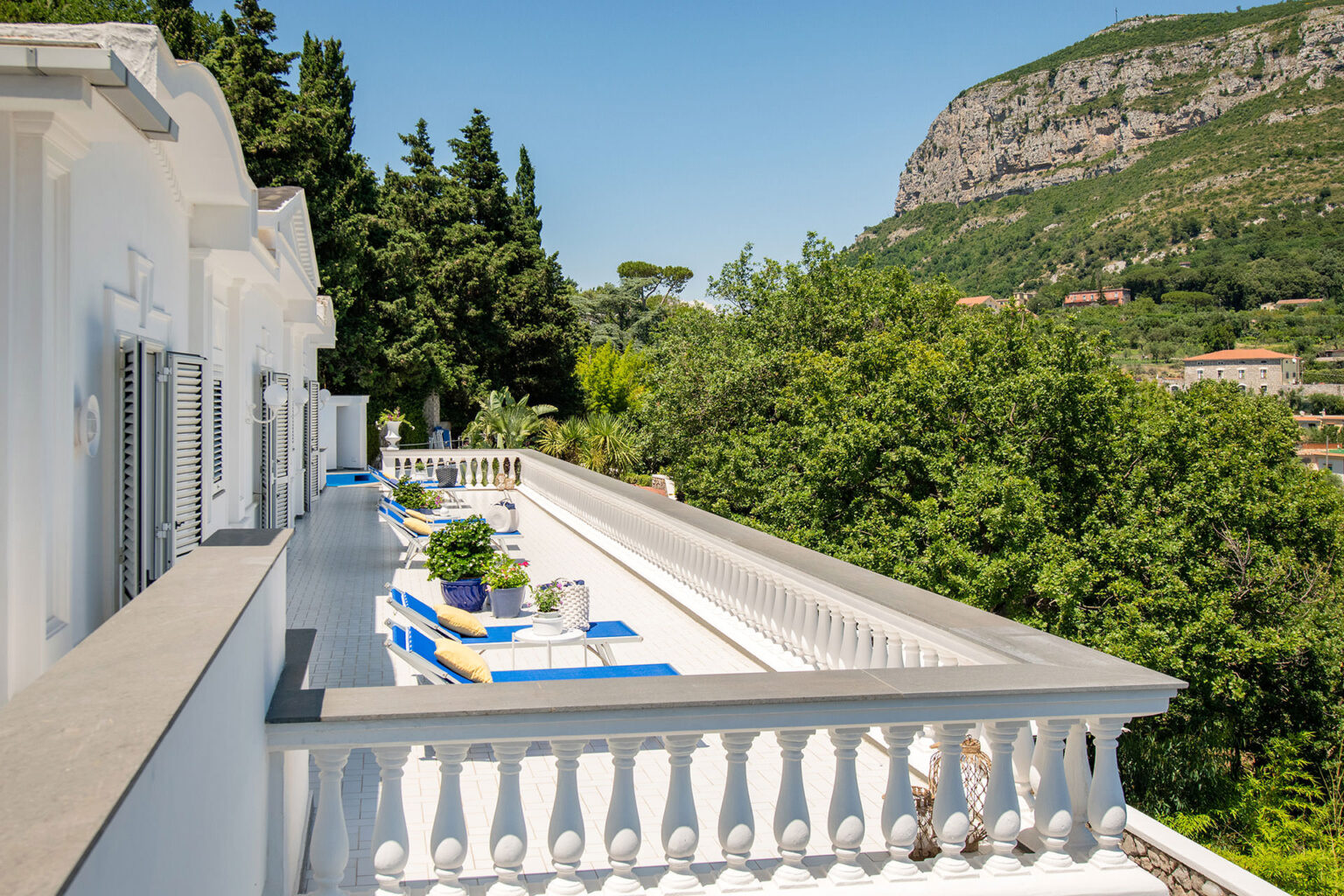 Scenic balcony view overlooking lush greenery and mountains, featuring blue lounge chairs and potted plants, ideal for relaxation and outdoor enjoyment. | Only on SopranoVillas