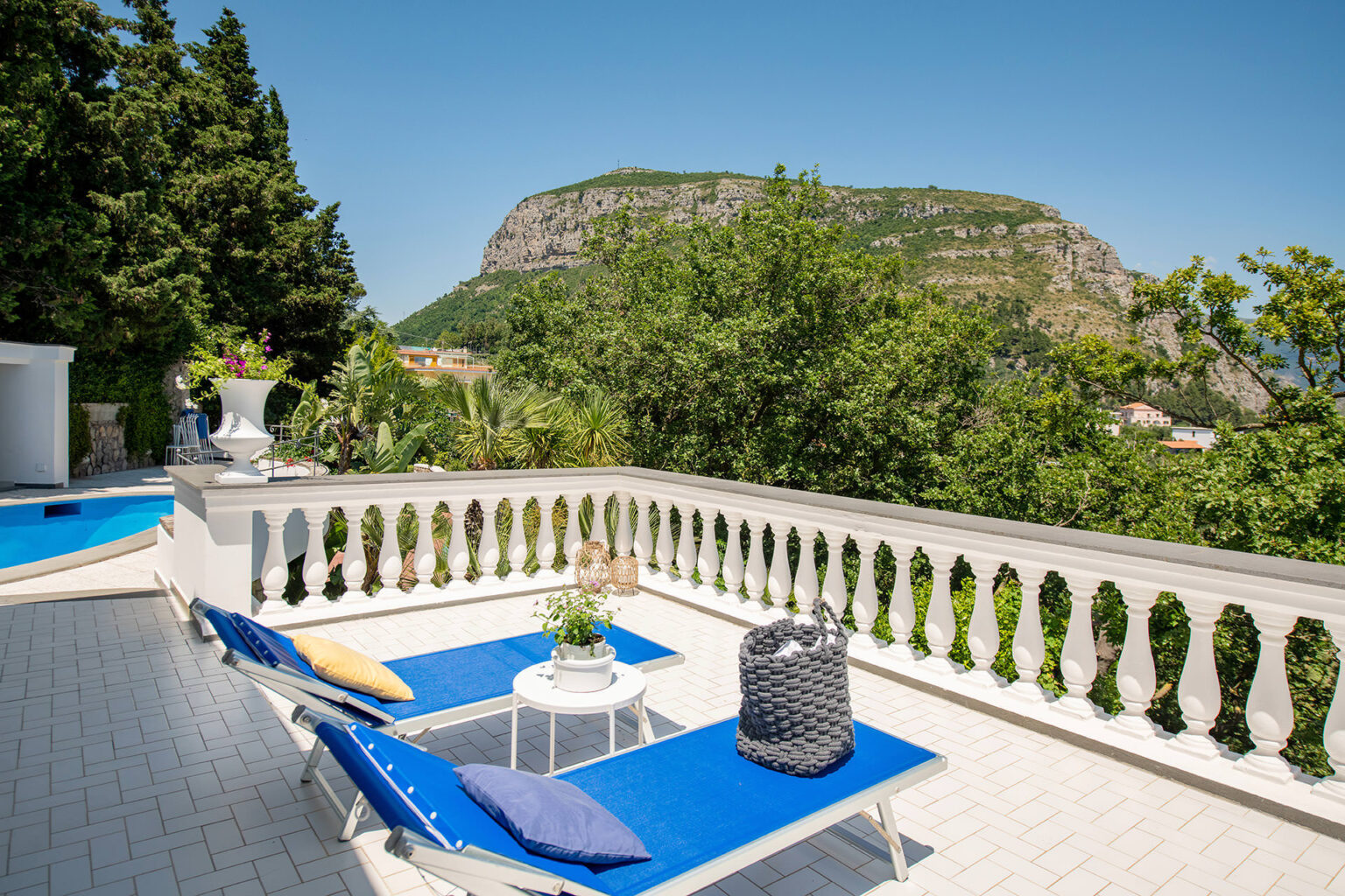 Sunny terrace with blue lounge chairs overlooking a scenic hillside, featuring greenery and a swimming pool, perfect for relaxation and outdoor enjoyment. | Only on SopranoVillas