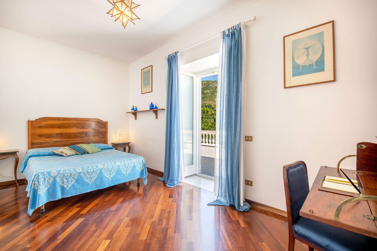 Bright and airy bedroom featuring a wooden bed with blue bedding, hardwood floors, and large windows with blue curtains. The room includes a wooden desk and decorative wall art, creating a cozy and inviting atmosphere. | Only on SopranoVillas