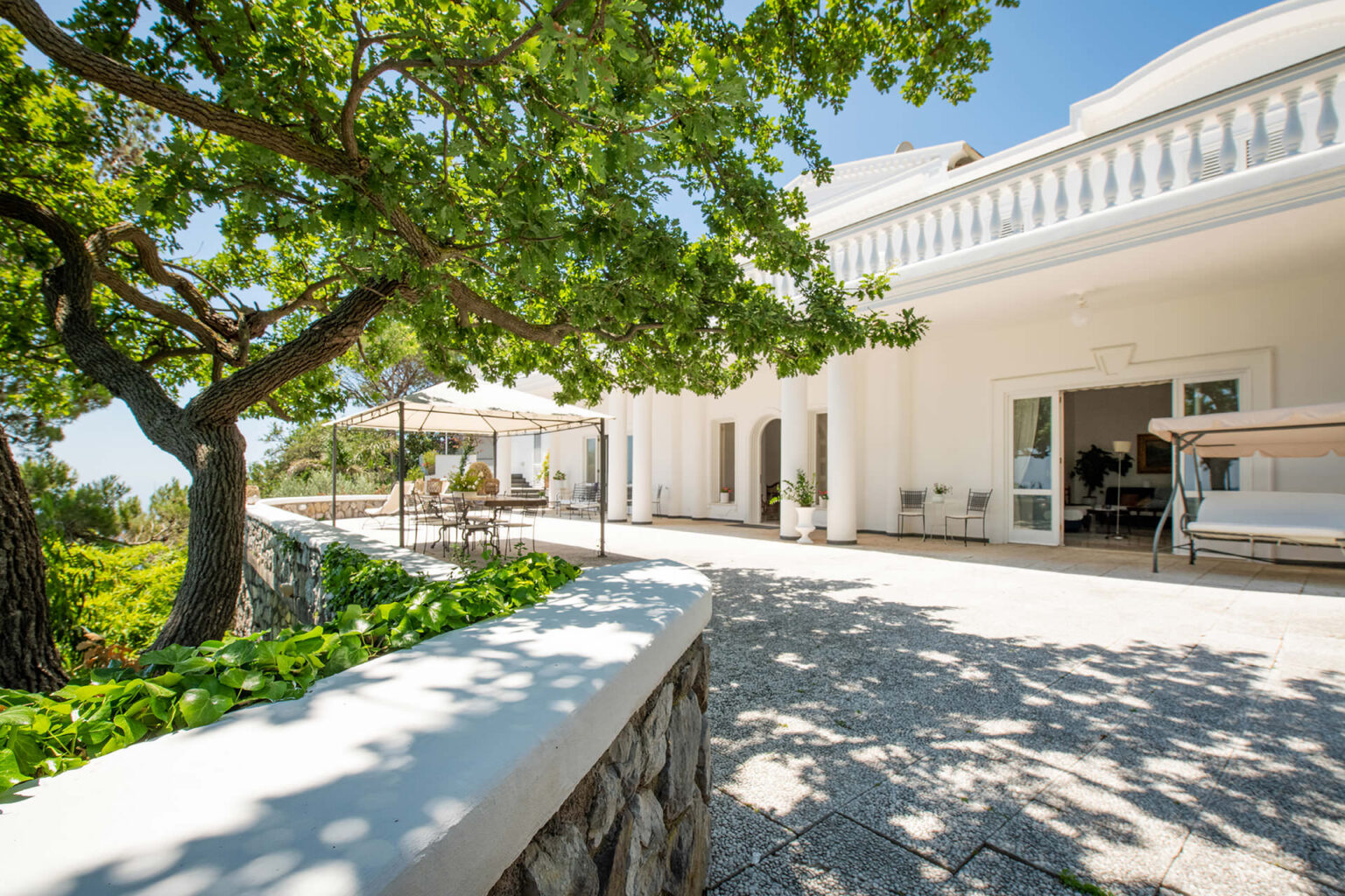 Luxurious villa exterior featuring a spacious terrace surrounded by lush greenery, a large tree, and outdoor seating under a canopy, perfect for relaxation and entertaining. | Only on SopranoVillas