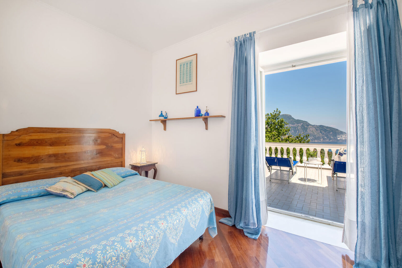 Cozy bedroom with a blue bedspread, wooden headboard, and a view of the mountains and outdoor seating area through an open balcony door. Natural light floods the room, complemented by blue curtains and minimalist decor. Ideal for a relaxing getaway. | Only on SopranoVillas