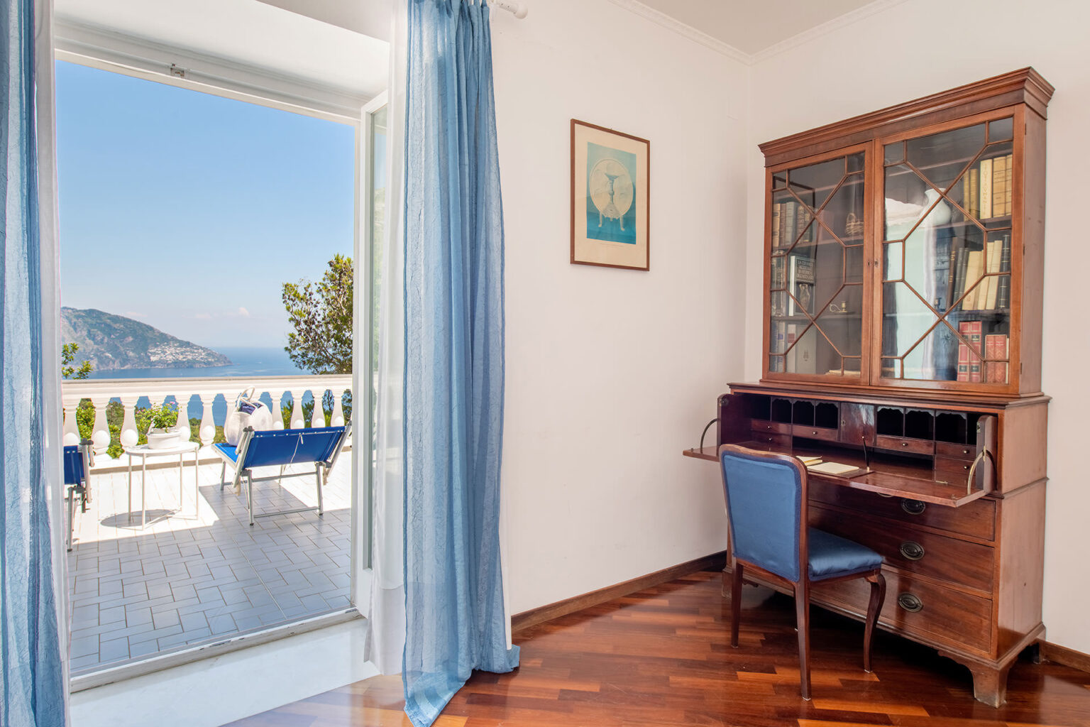Bright and airy interior featuring a wooden writing desk and chair, with a view of a scenic outdoor patio and mountains through an open balcony door. Elegant blue curtains frame the entrance, enhancing the tranquil atmosphere. | Only on SopranoVillas