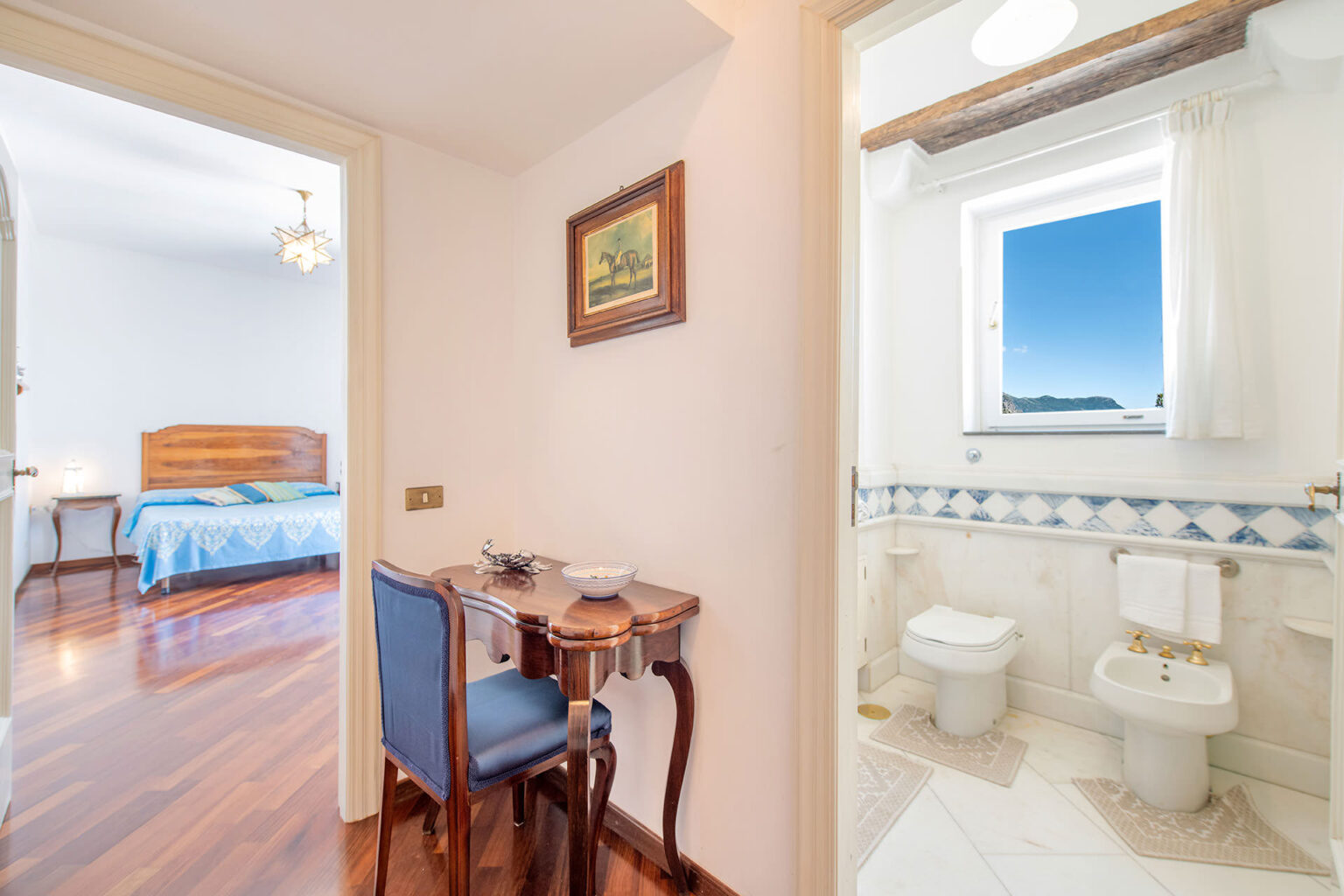 Interior view of a cozy bedroom with a blue bedspread and a small study area featuring a wooden desk and chair, adjacent to a well-lit bathroom with modern fixtures and a window offering a scenic view. | Only on SopranoVillas