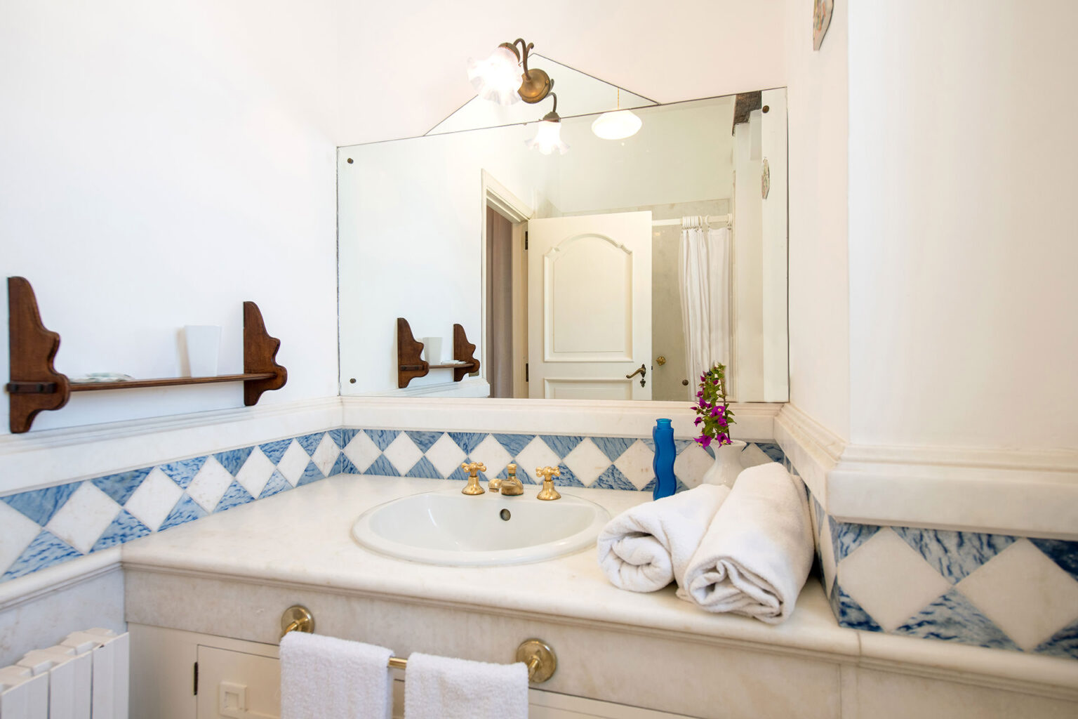 Modern bathroom interior featuring a white marble countertop with a sink, golden faucets, and neatly rolled white towels. The walls are adorned with blue and white tiled accents, complemented by wooden shelves. A large mirror reflects the space, and a small flower vase adds a touch of decor. Soft lighting enhances the inviting atmosphere. | Only on SopranoVillas
